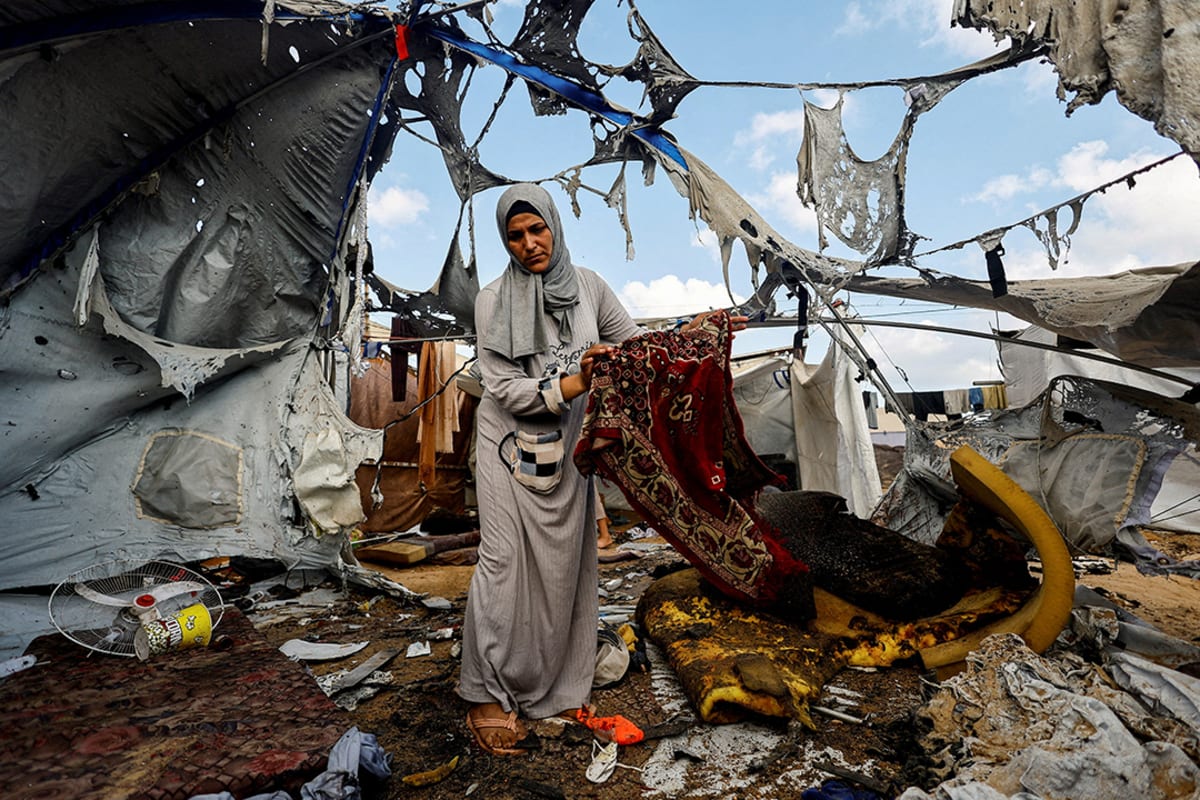 A woman stands amid the wreckage of a tent holding a prayer mat.