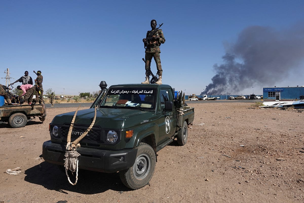A soldier stands on top of a truck holding a gun while black smoke billows in the distance.