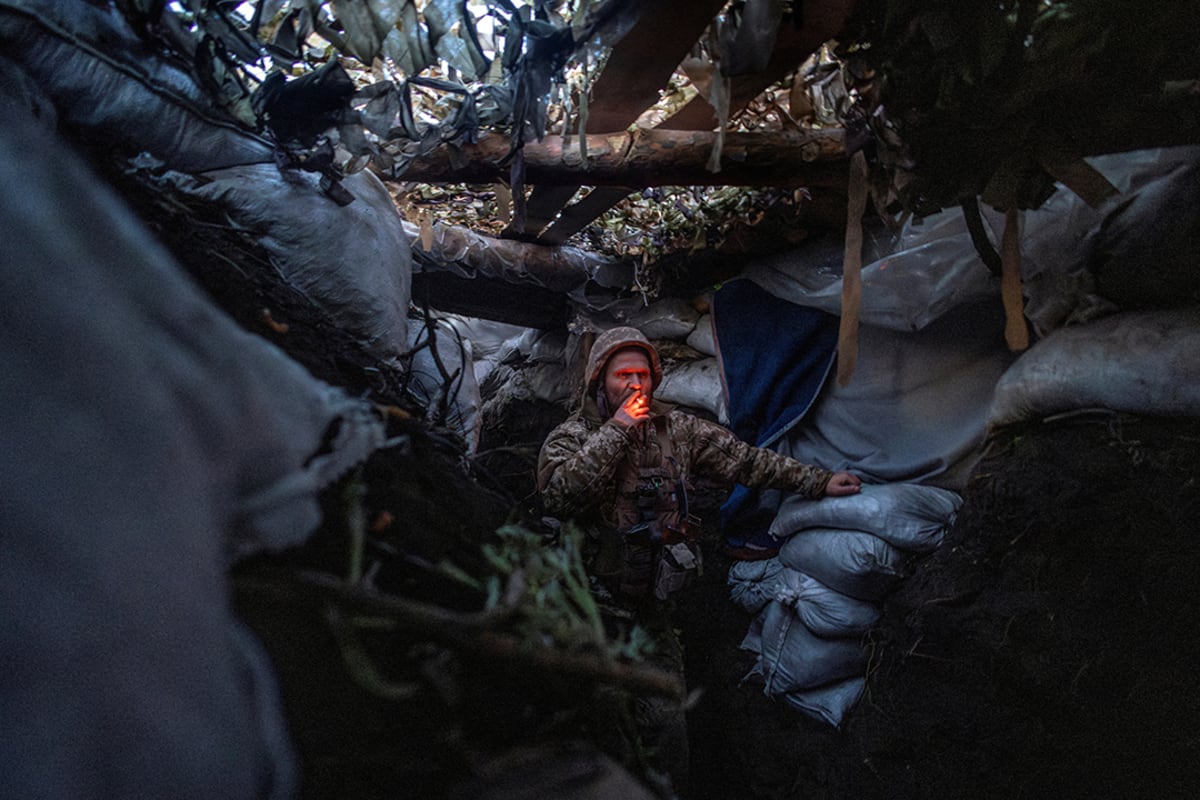 A soldier hunches in a trench while smoking a cigarette.