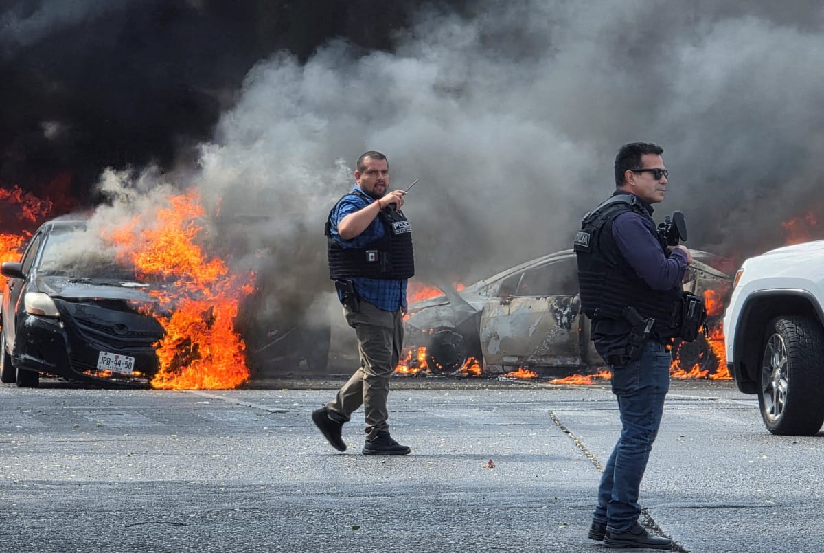 Two men stand in front of burning cars