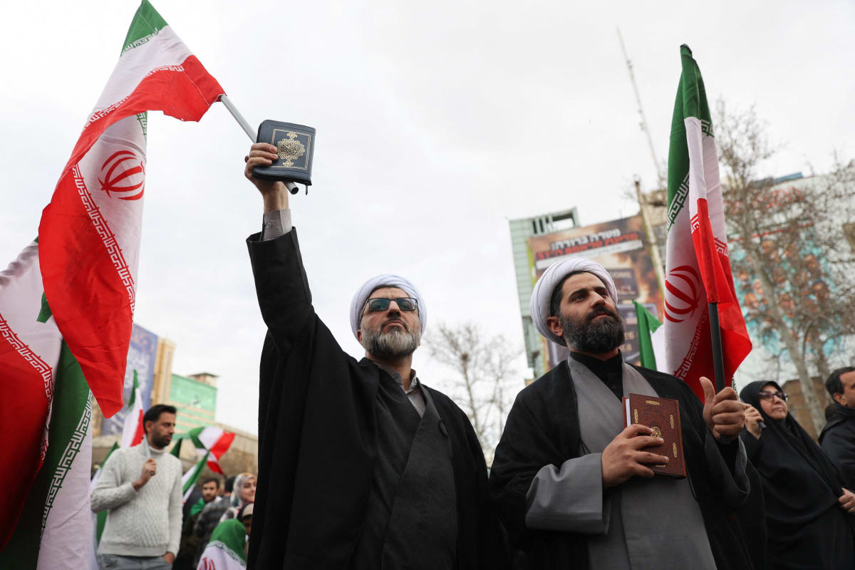 Two men in clerical clothes hold religious books in their hands and wave Iranian flags.