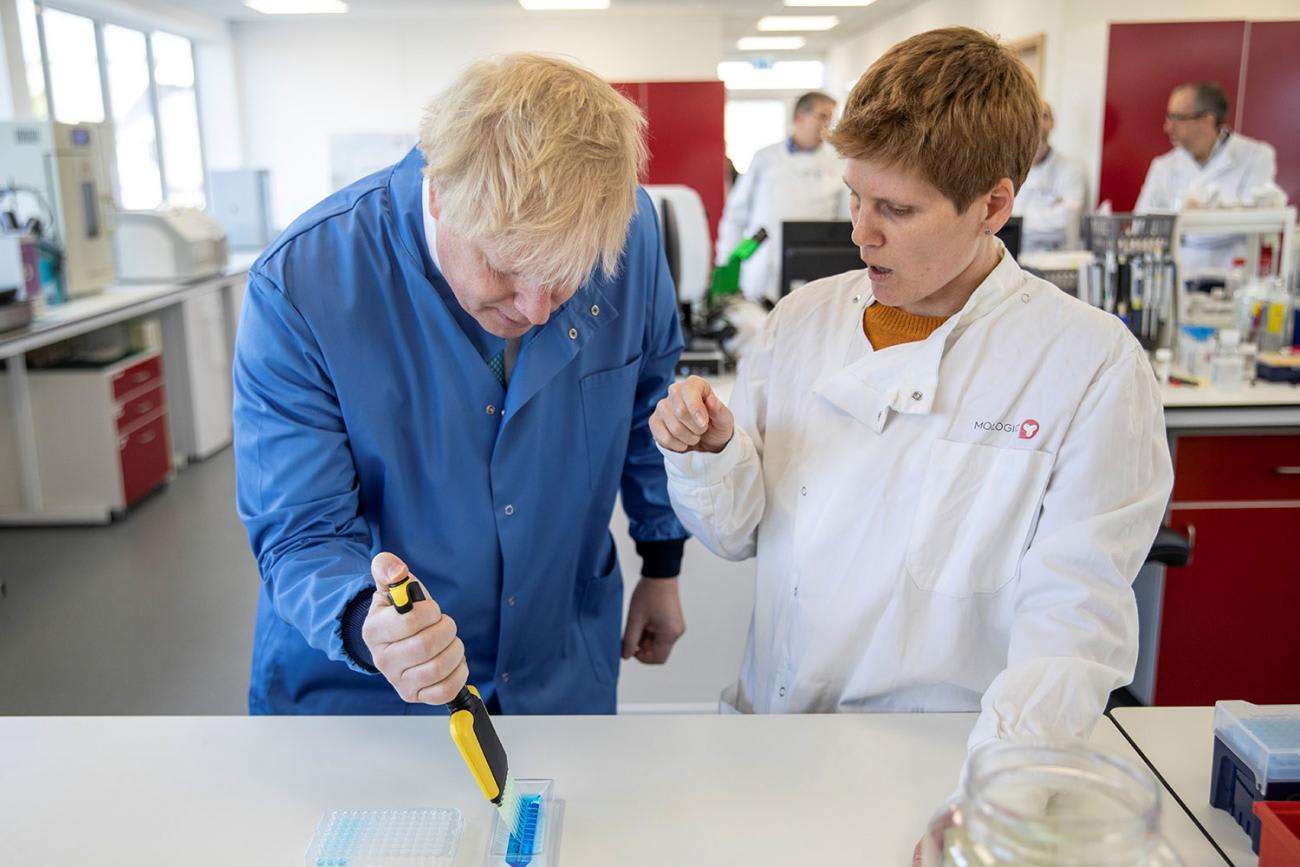 Photo shows the prime minister leaning over a laboratory bench, looking down, and carefully pipetting into a vessel while wearing gloves. A scientist stands to his left speaking to him.