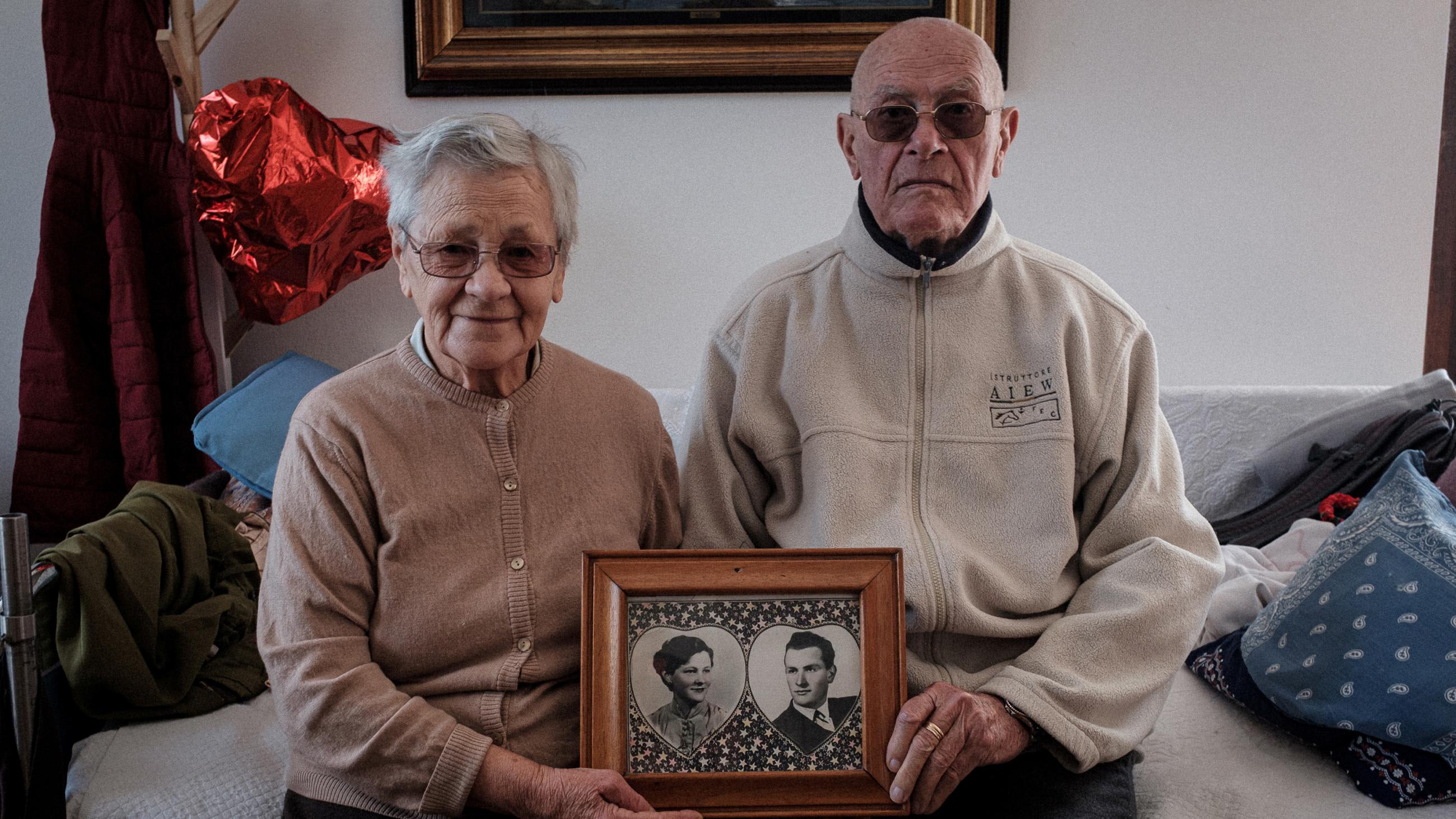 Picture shows the elderly couple sitting together holding a picture of the two of them taken many years before. They are a cute couple.