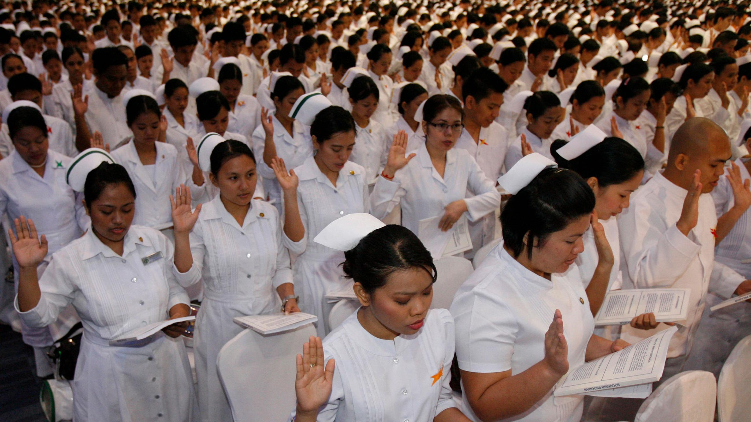 Picture shows thousands and thousands of nurses in uniform lined up with right hands raised reading from a thick packet. The women have traditional nurse caps, but the men's heads are bare.