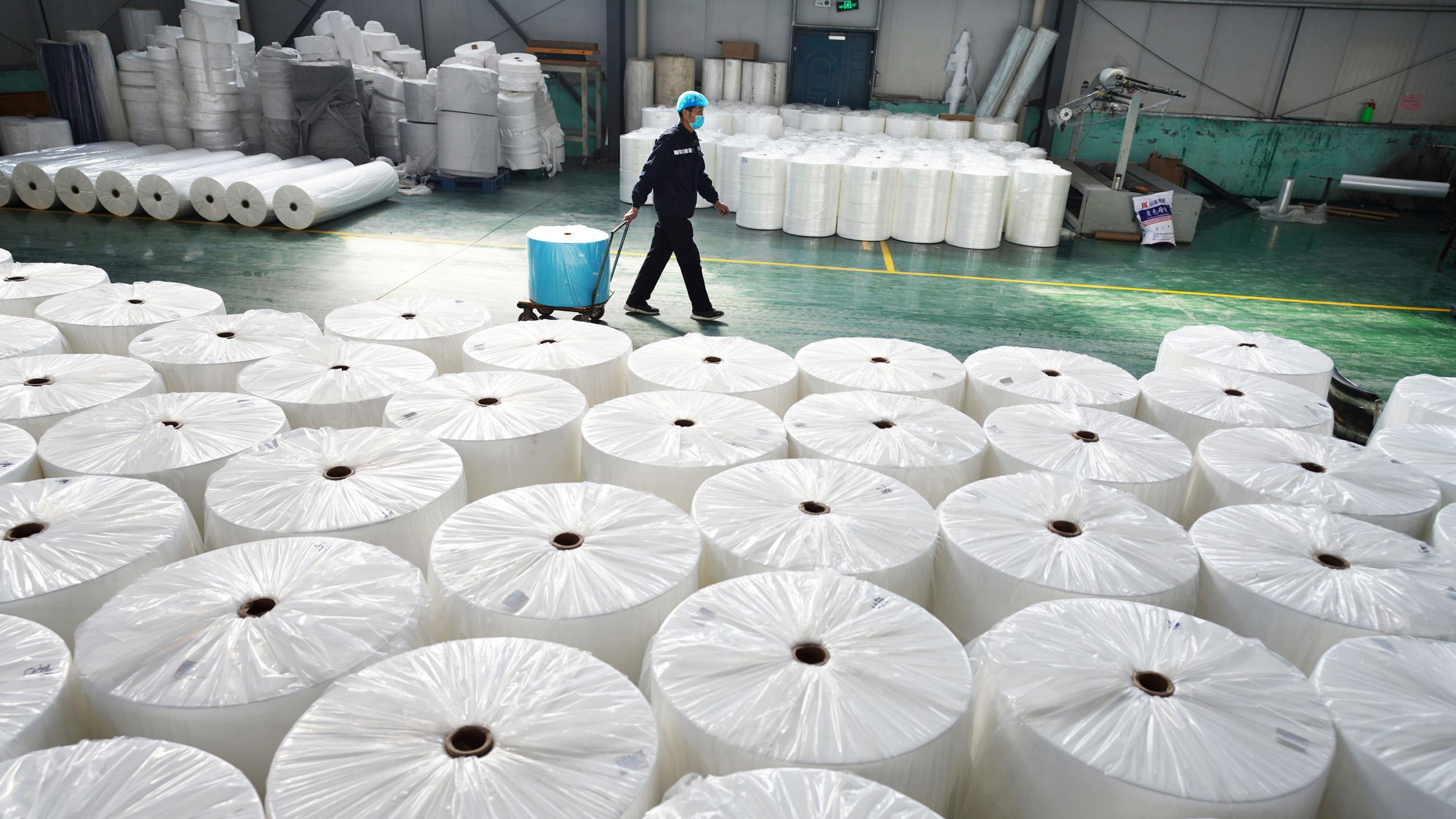 Picture shows a large factory space with a massive stack of large fabric spools in the foreground and a single worker waling by in the background.