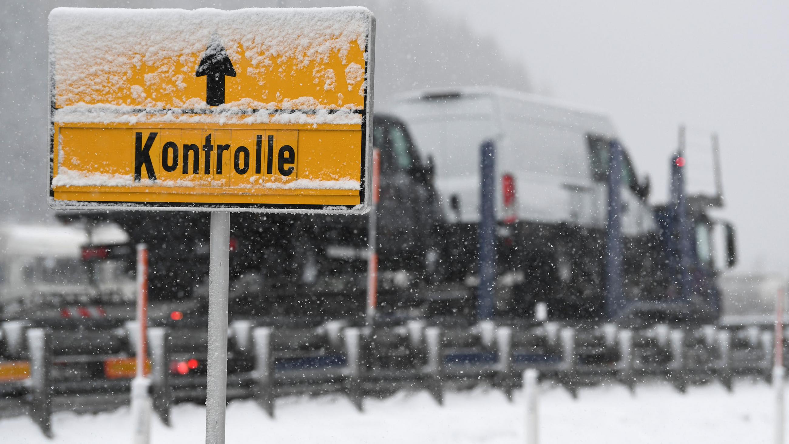 Picture shows a yellow sign and trucks waiting at the checkpoint. Snow is falling and partially obscures the sign.