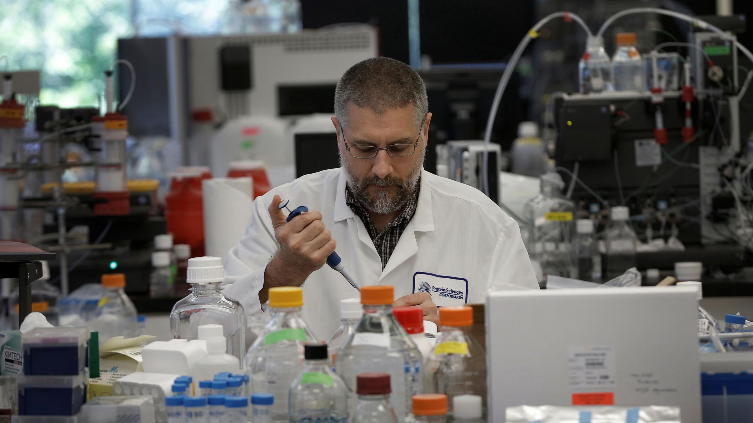 Picture shows the researcher pipetting into vials in a laboratory surrounded by chemical bottles and instruments.