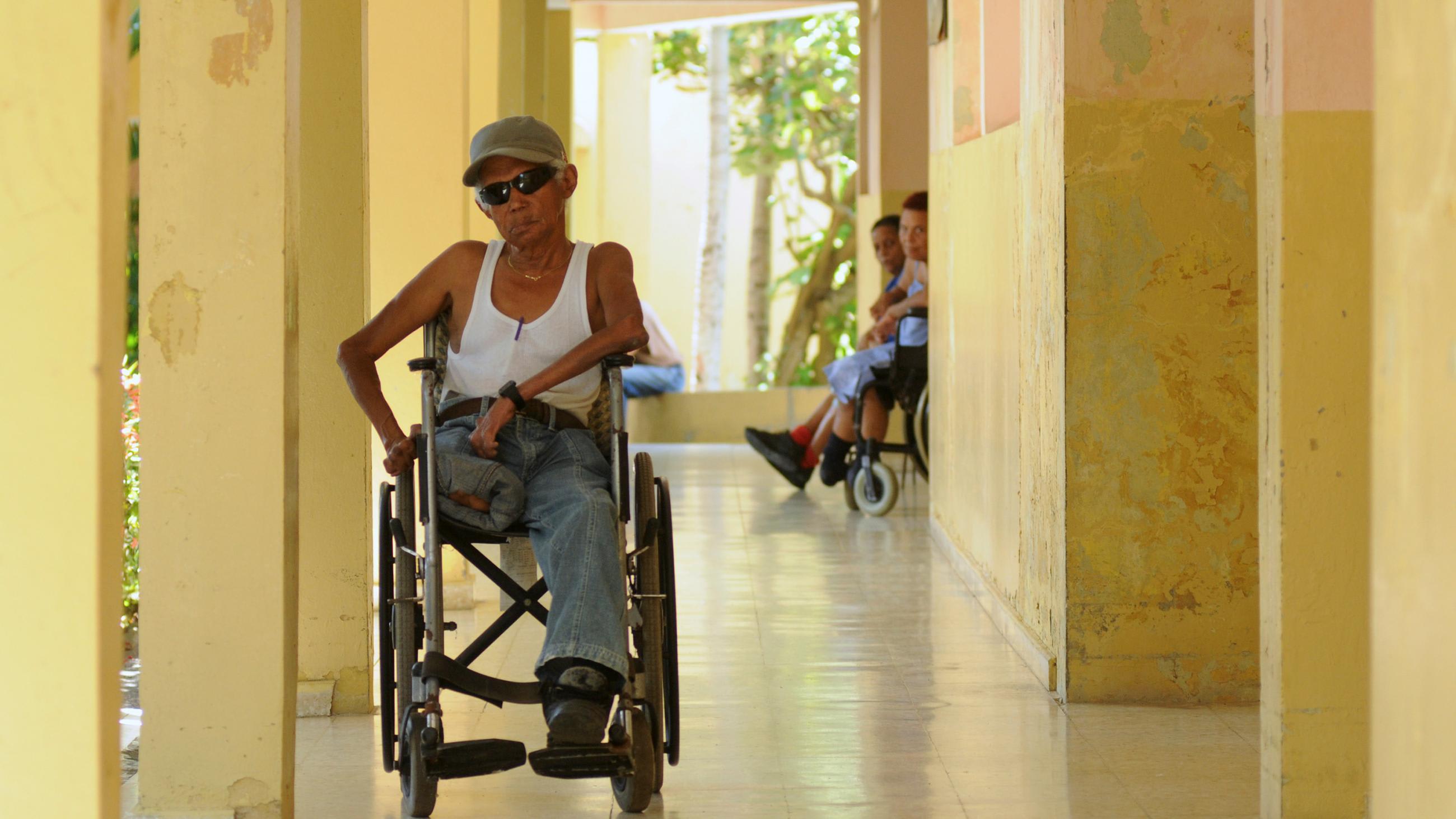Picture shows Domingo in a wheelchair wearing dark shades and a white tank top. It appears to be a warm summer day.