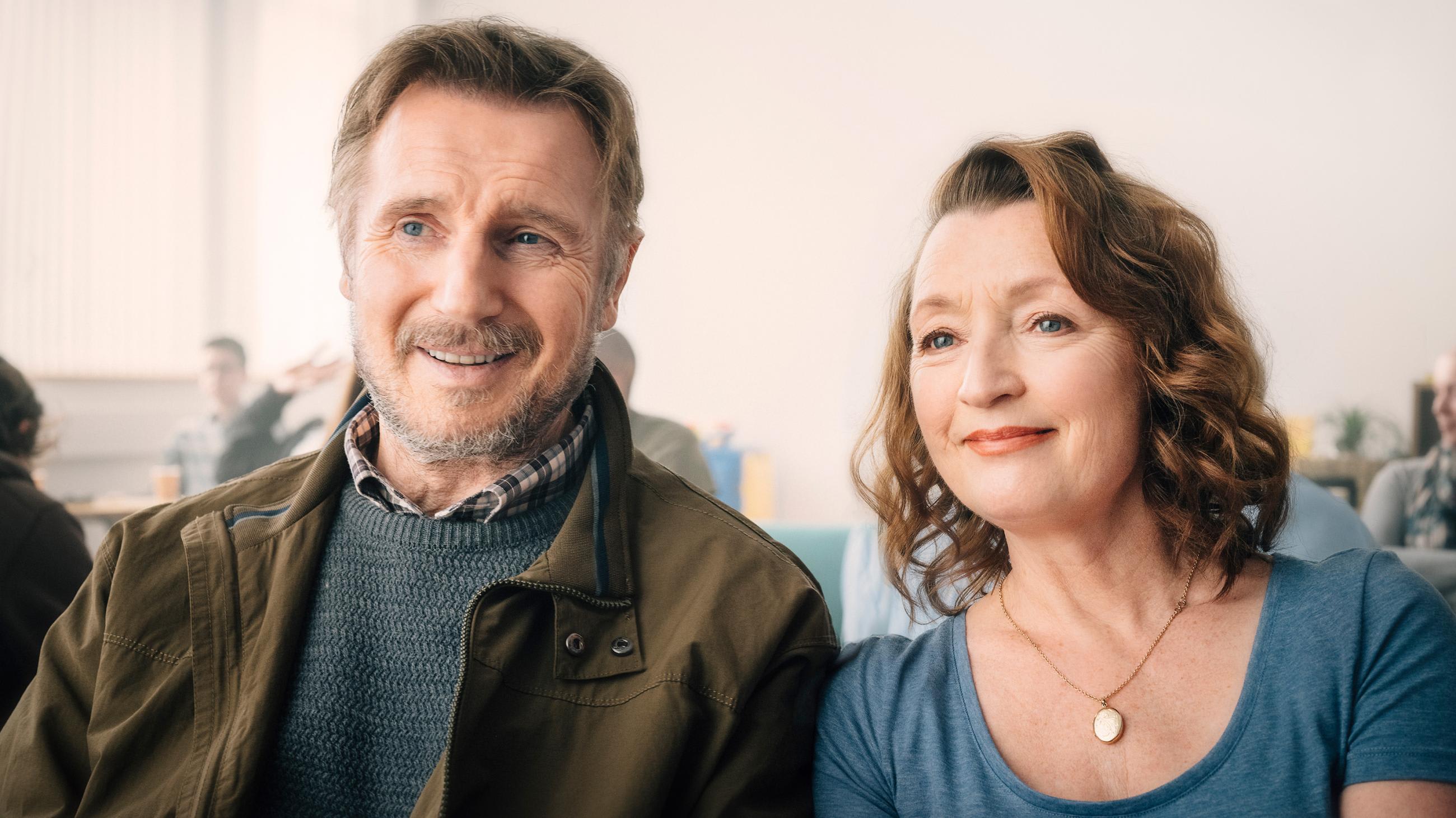 Image is of the two actors sitting together and smiling in a hospital waiting room.