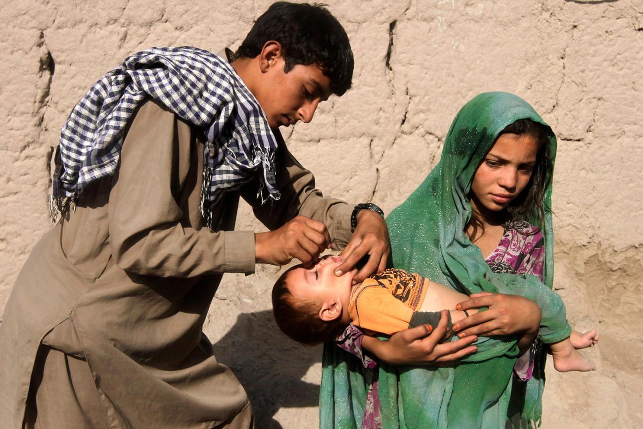 A child receives polio vaccination during an anti-polio campaign on the outskirts of Jalalabad, Afghanistan on August 18, 2014. In the photo a young man with a black-and-white checkered scarf slung over his shoulder administers drops to a baby held in the arms of a young woman wearing a bright green headdress.