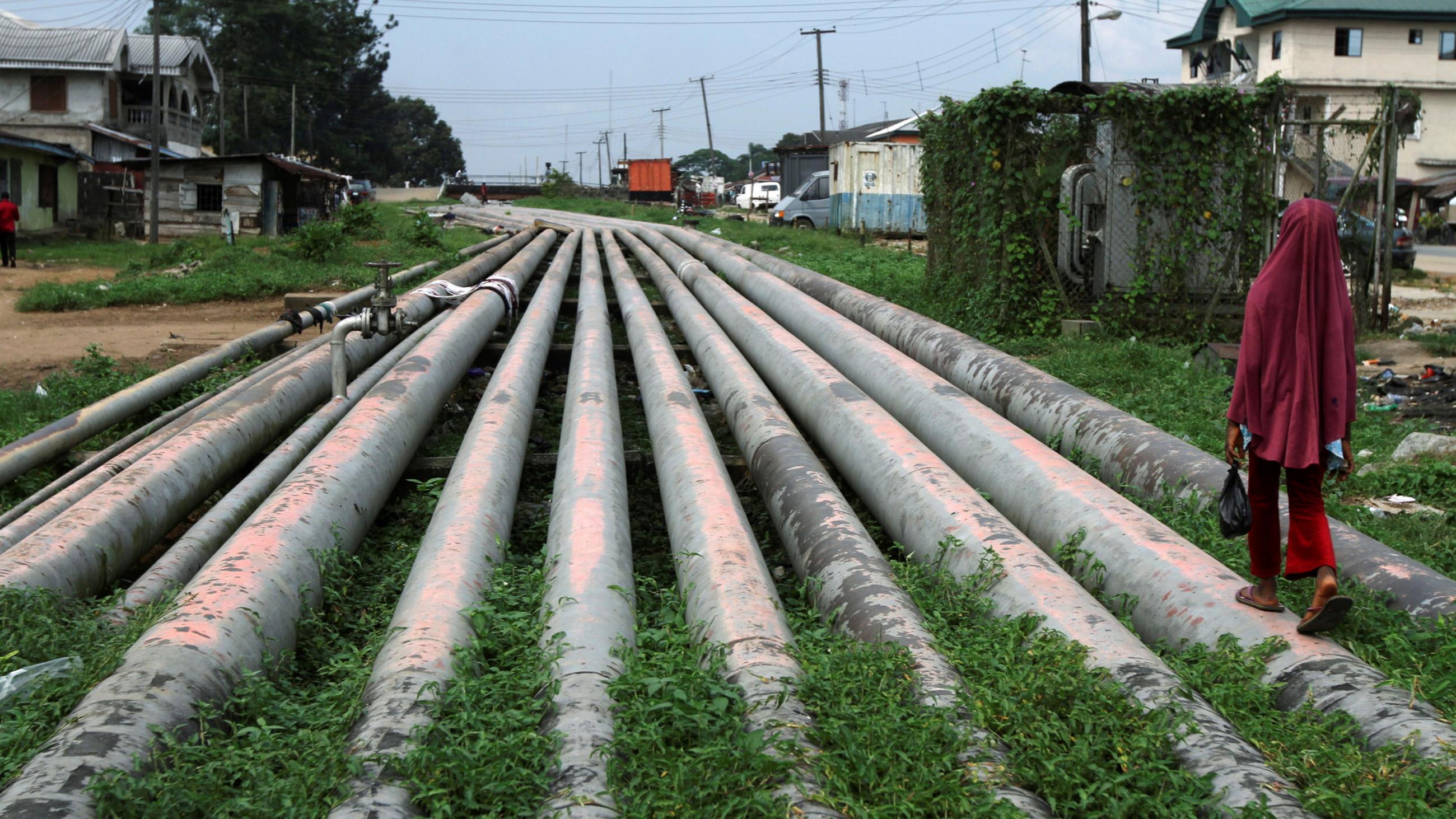 This is a dramatic picture of a dozen or so large pipes, each a couple feet in diameter, running parallel through what appears to be a rural area. The pipes extend off into the distance, forming a natural vanishing point, and a girl wearing red is walking along one of the pipes with her back to the camera.