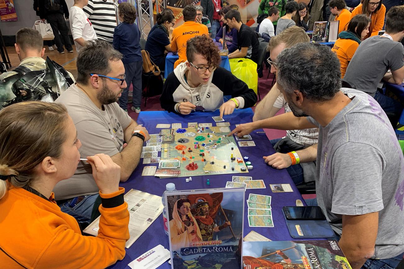 Photo shows five players at long table avidly discussing the situation on the game board, which shows a map of Western Europe. Spread around the table are cards and colorful game pieces. Lots of other tables are in the background with similar games going on.