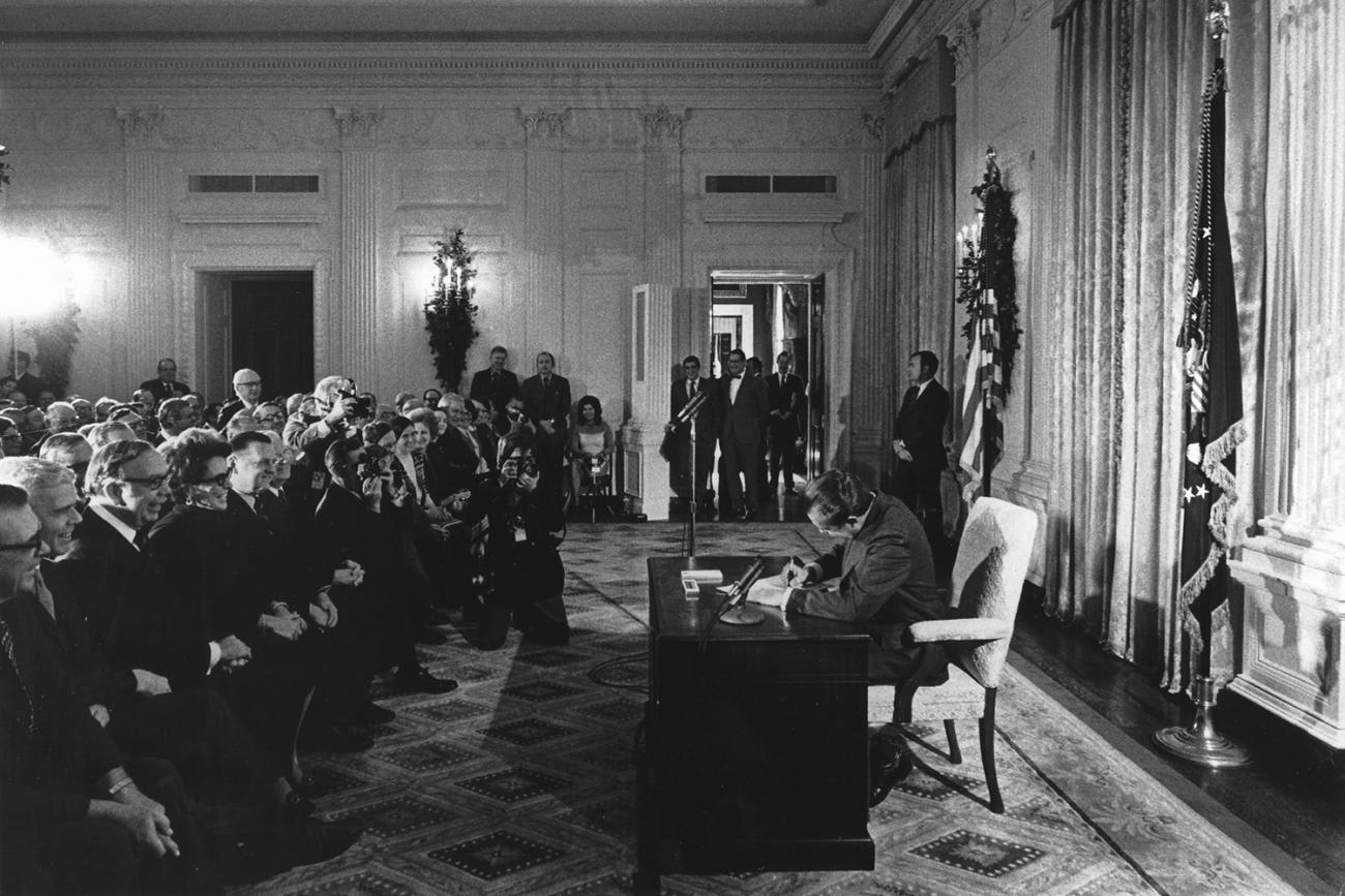 The image shows Nixon at his desk, his head down, signing the bill. In front of him is a large audience of people, many of whom are smiling broadly.