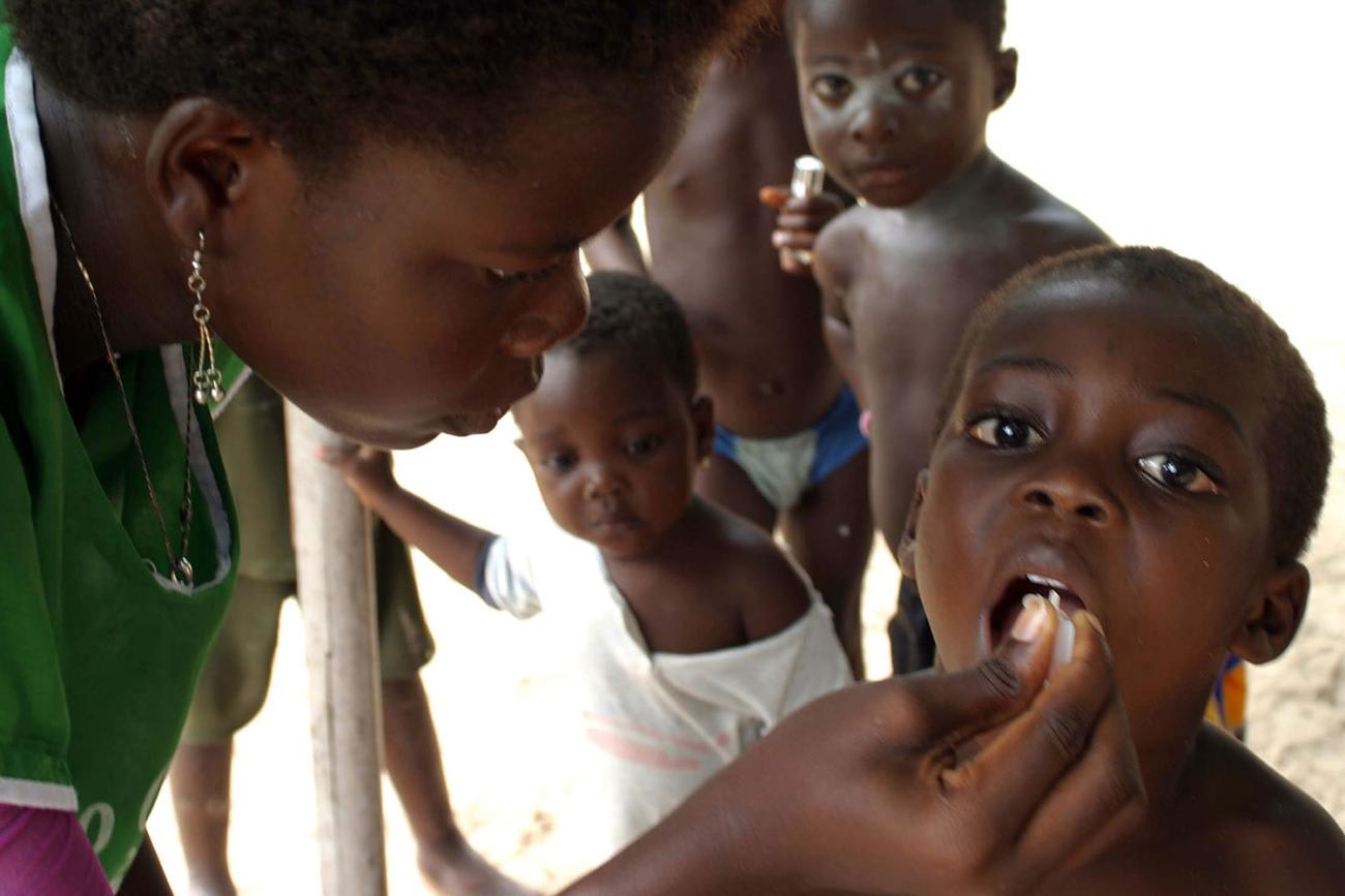 Picture shows a health care worker in a green uniform giving oral drops to a young boy who has his mouth open. Several other young children can be seen queued behind the boy.