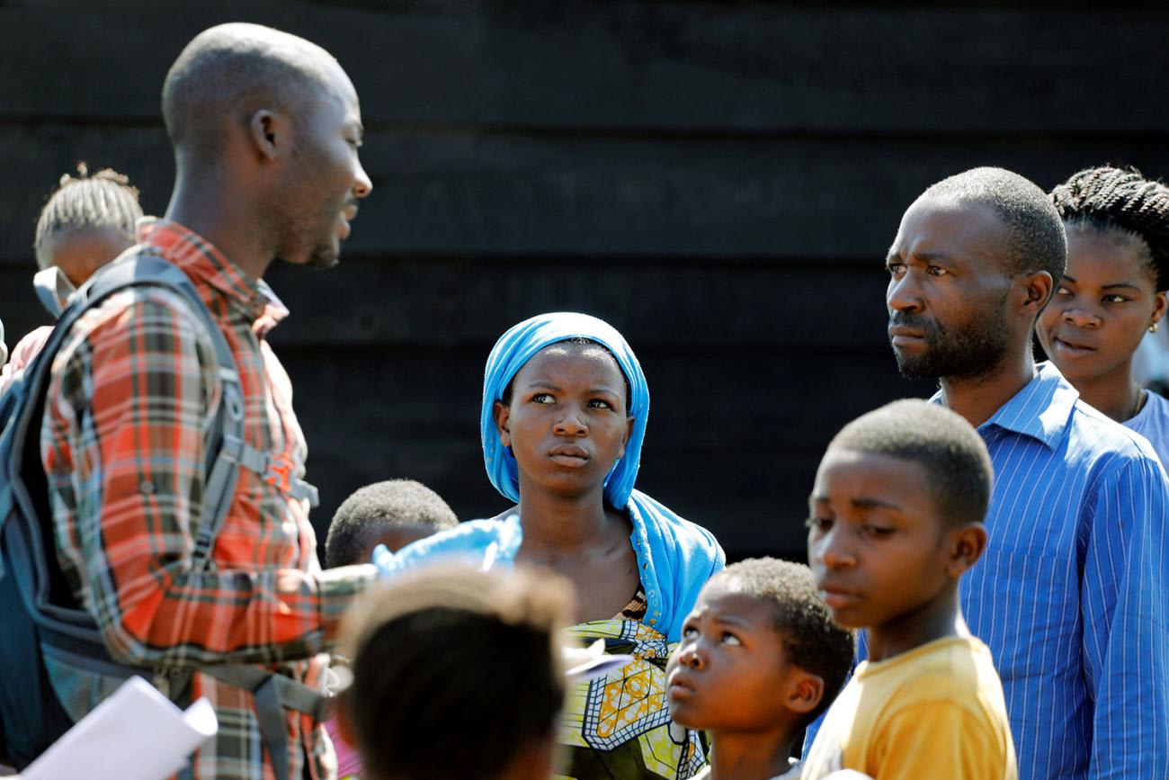 The picture shows a small group gathered around a health care worker who is wearing an orange shirt and carrying a blue backpack. The crowd is mostly children, but a few adults with serious, scrutinizing looks on their faces can be seen listening to the worker.