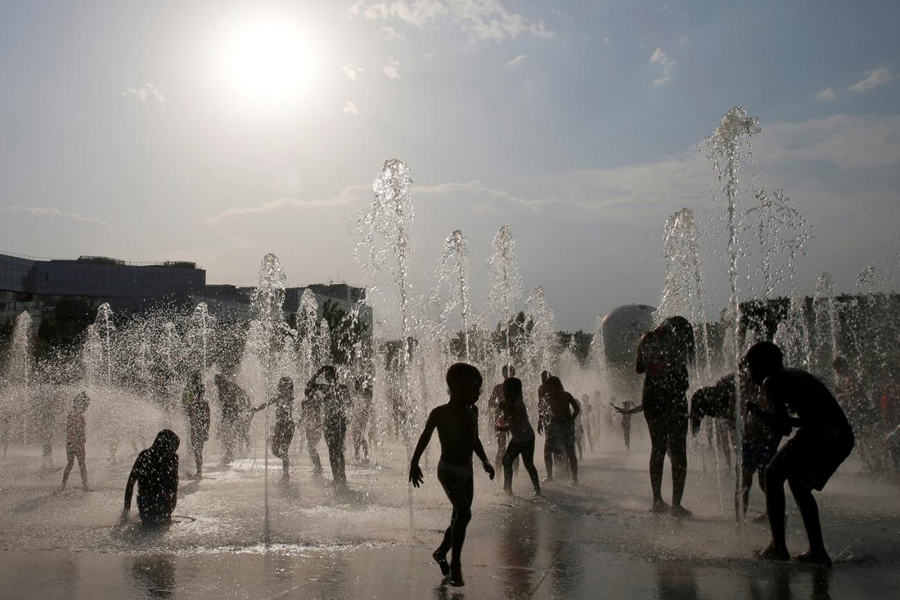 The picture shows a number of children silhouetted as they cool off in water fountains at an urban park.