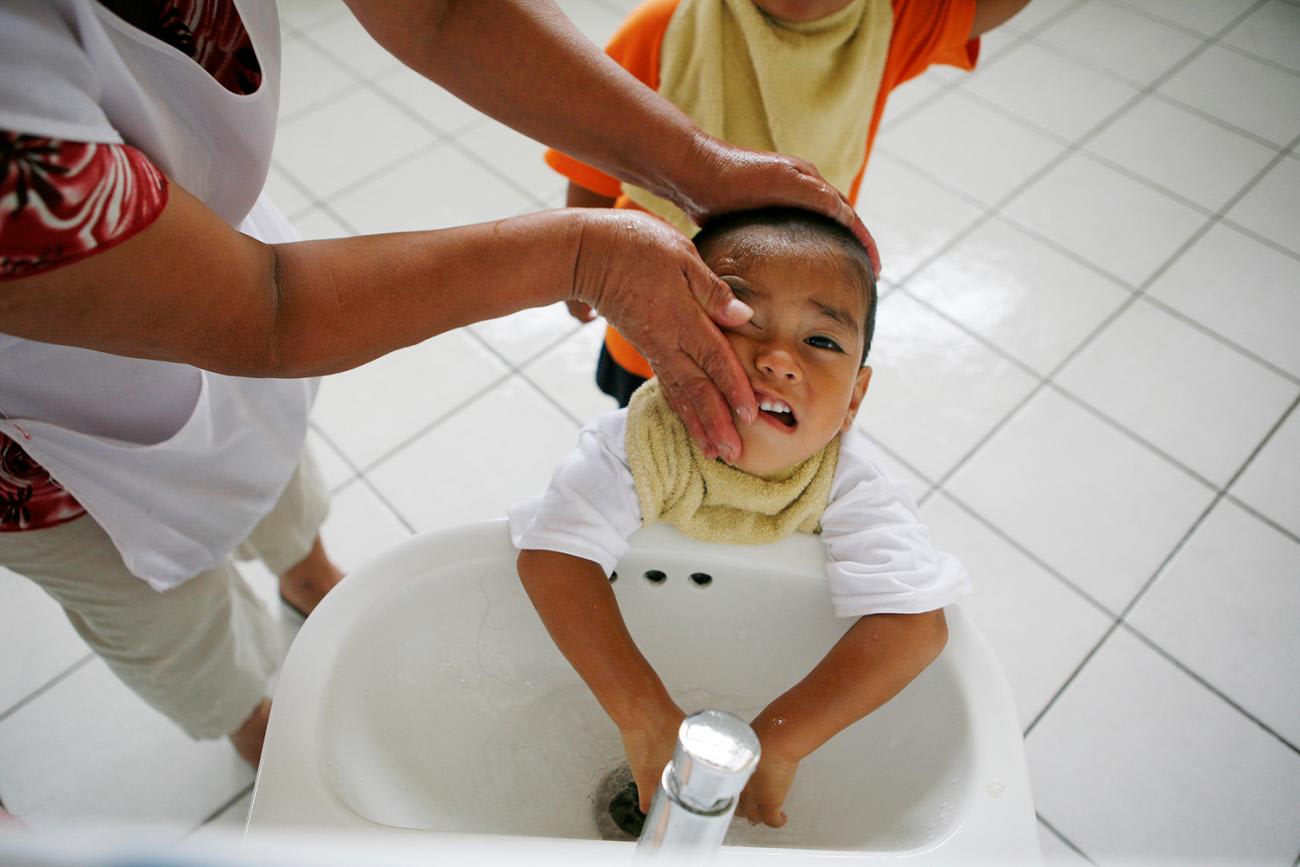 The picture shows a social worker washing the face of a child standing at the sink. He doesn't look like he's enjoying it.