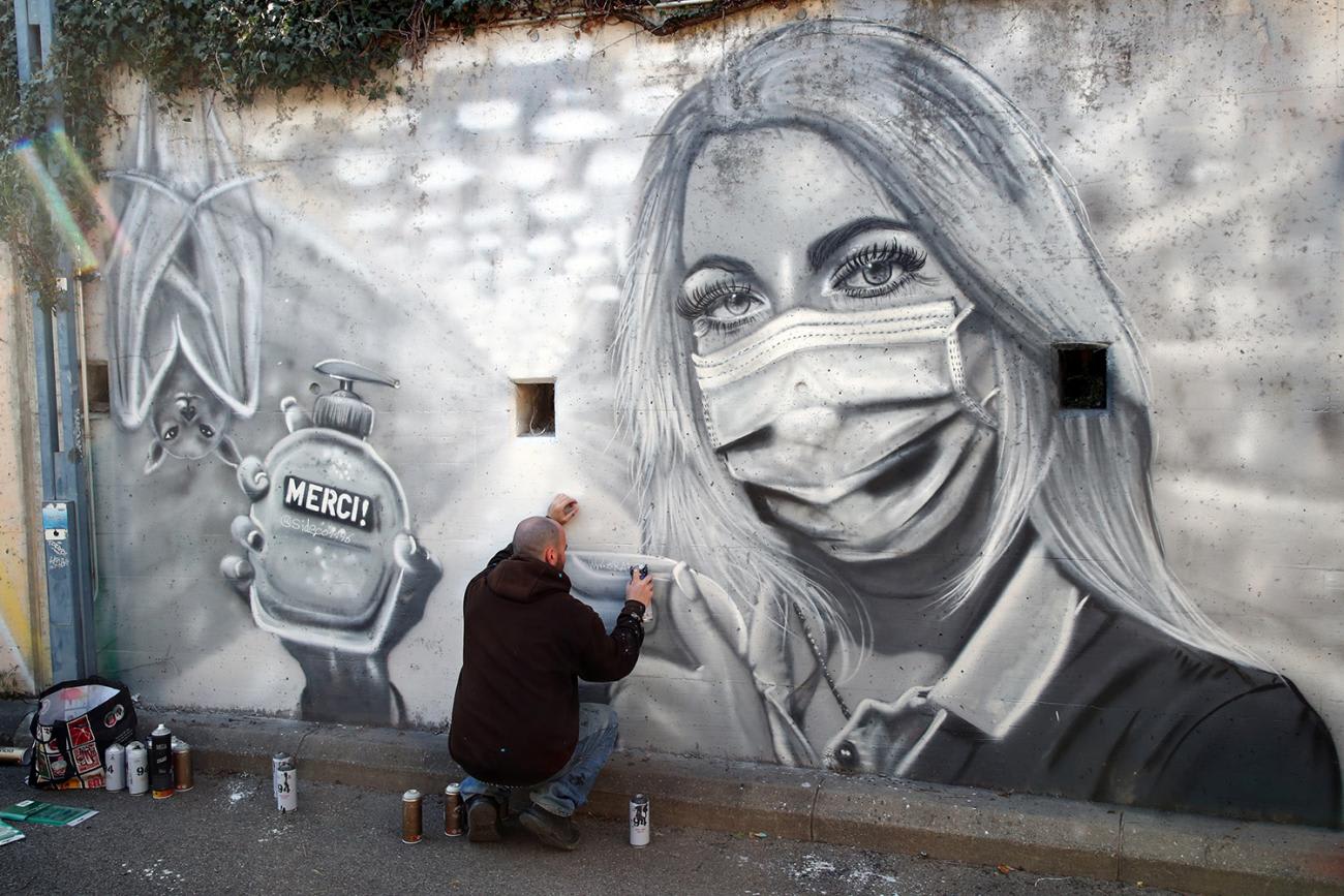 The photo shows the artist finishing the mural, which features a woman with a mask holding a bottle of hand sanitizer with the word merci (thank you) written on it.