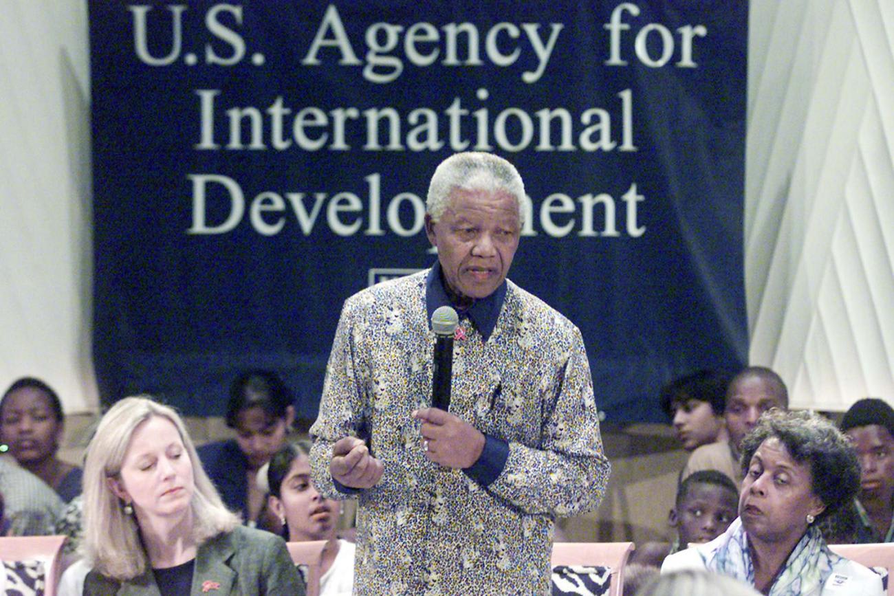 Image shows Nelson Mandela speaking into a microphone before a sign that reads, 