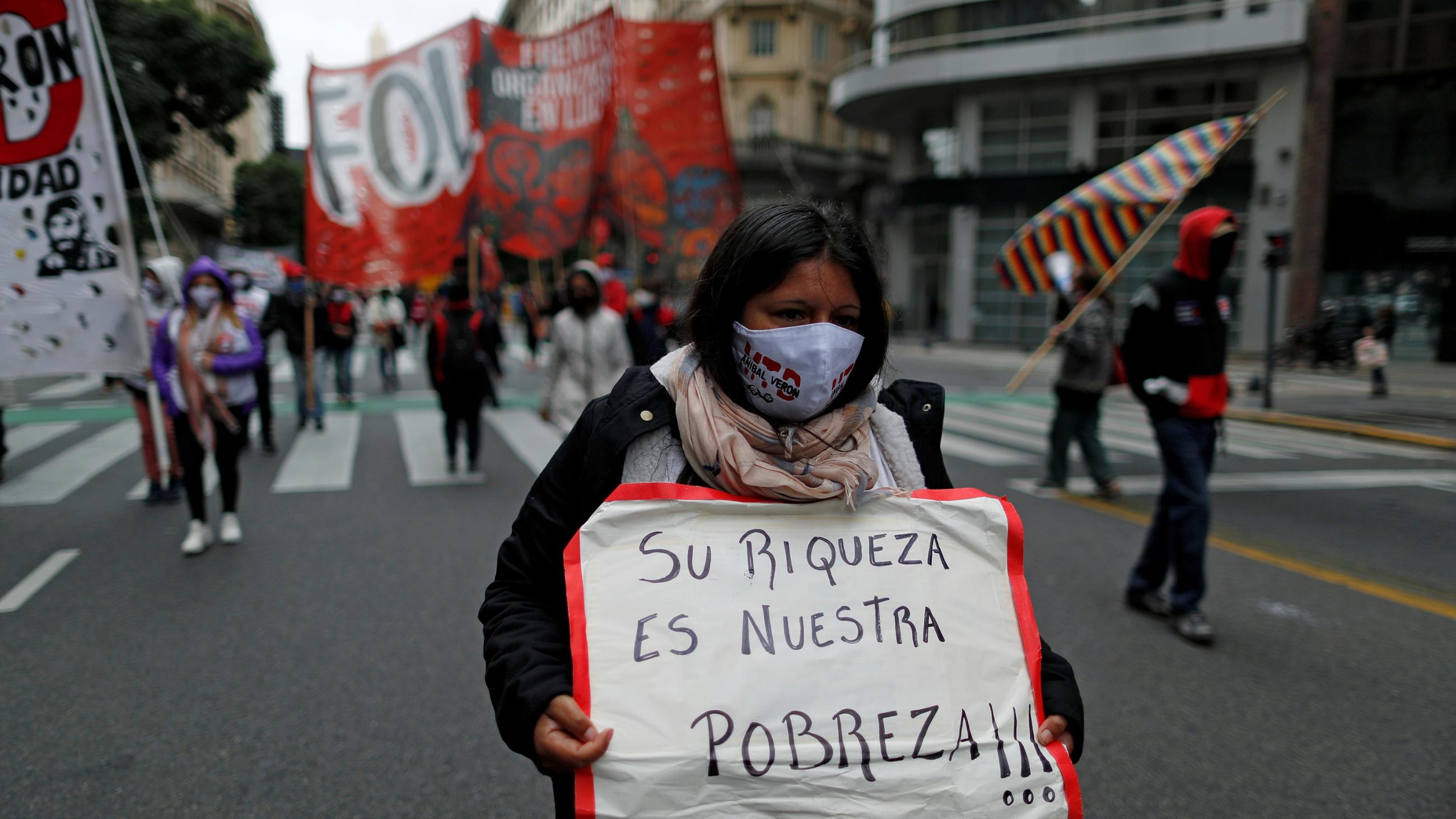 The photo shows a street protest with a woman in front holding a sign that reads "their richness is our poverty."