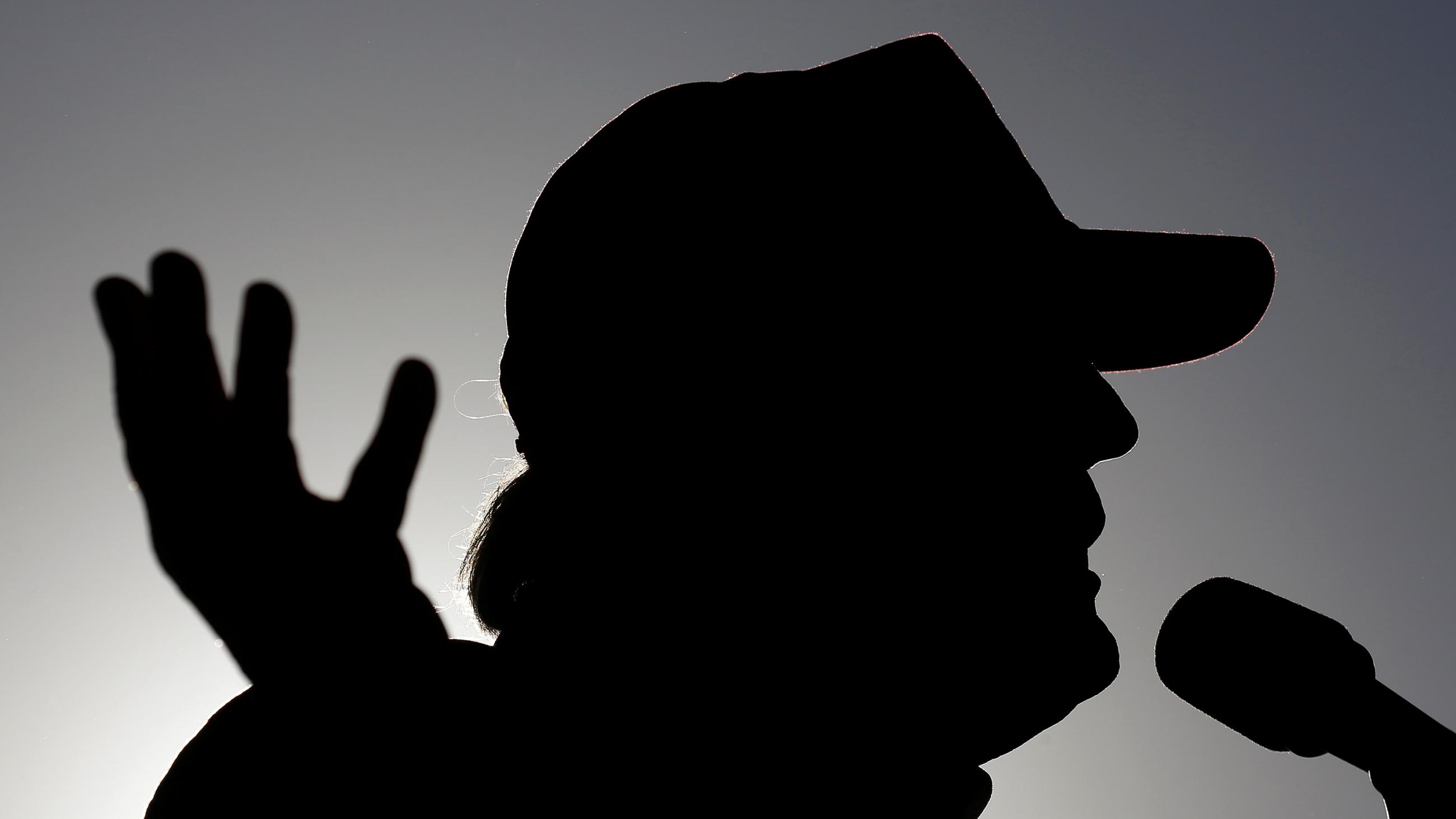 The photo shows a silhouette of the candidate speaking. he is wearing a hat and gesturing with his hand.