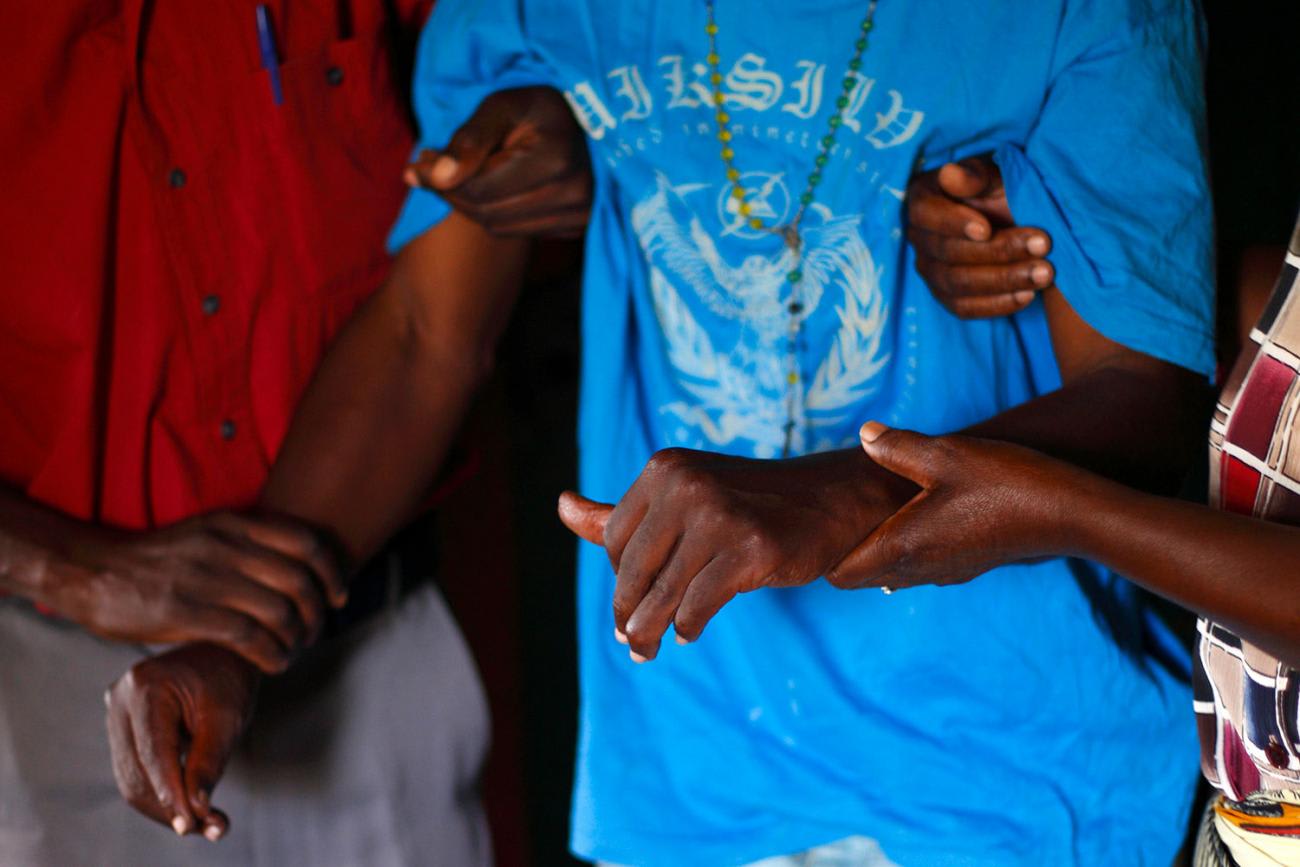 The picture shows the torso of a man wearing a loose-fitting light blue shirt being helped by two other people.