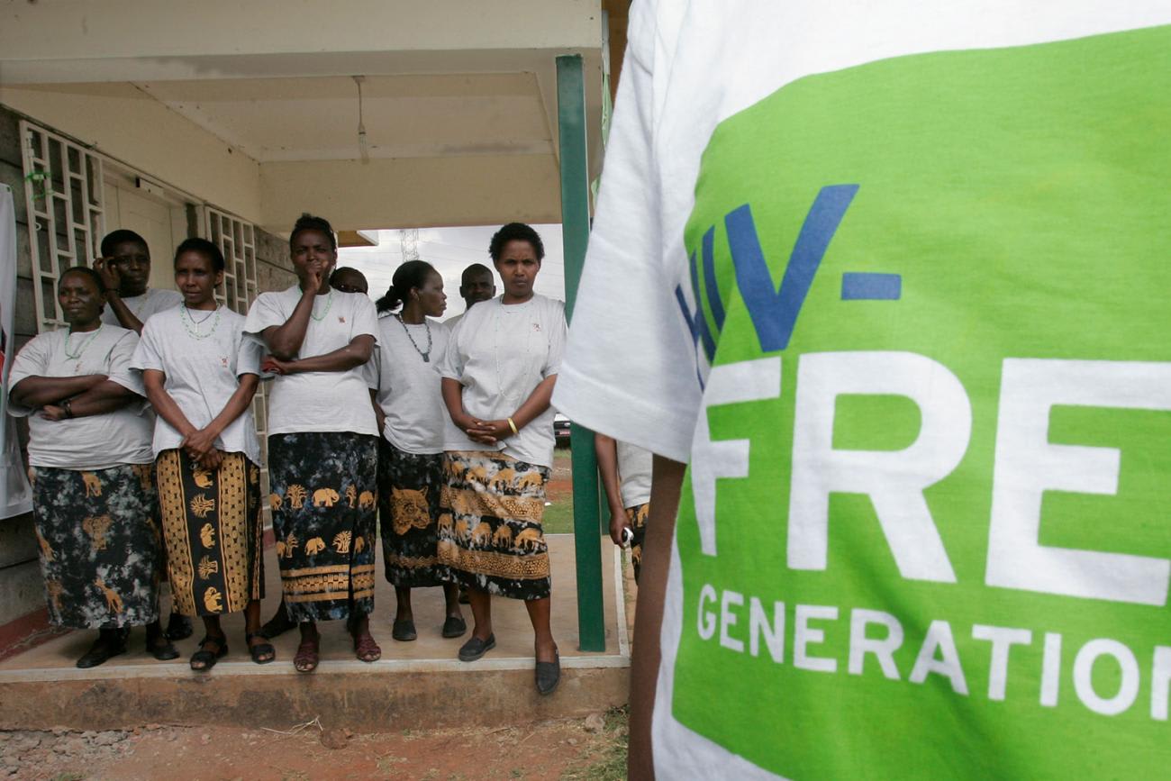 The photo shows several people about 20 feet from the camera standing and listening while a figure stands in the foreground. The camera is too close to see their face, but they are wearing a Tee Shirt that says, "HIV-free Generation."