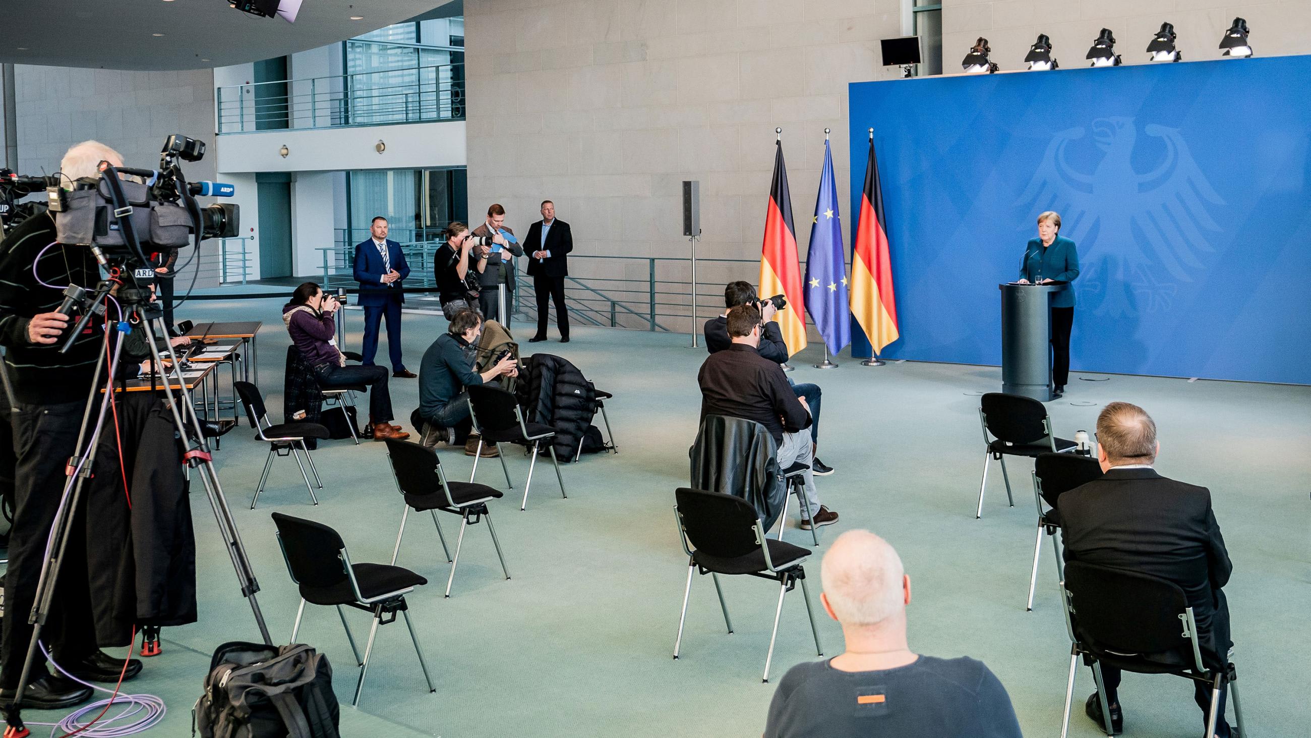 The photo shows the German Chancellor speaking to a room sparsely populated with reporters practicing social distancing.