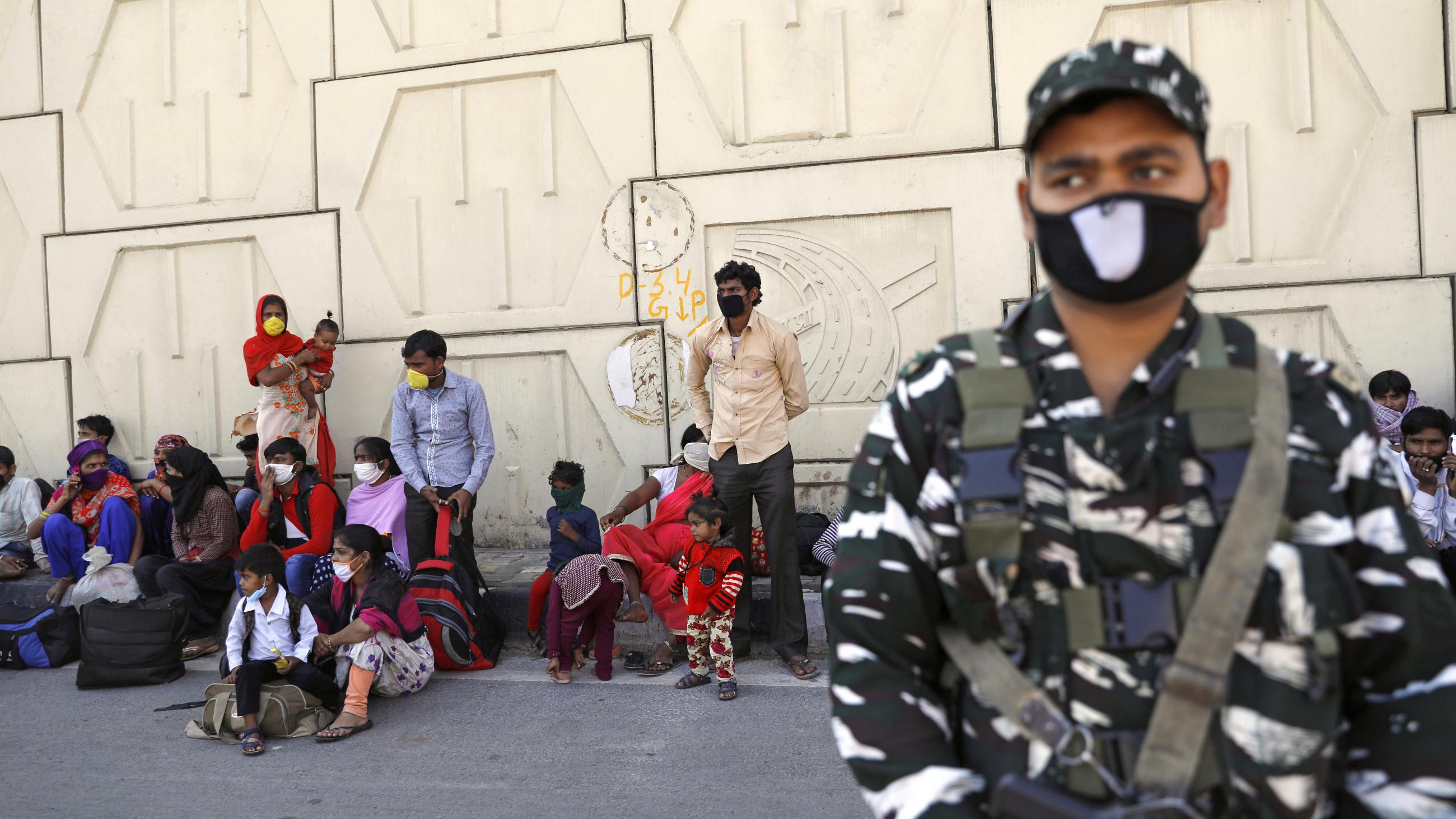 An Indian soldier in a camouflage uniform stands guard. Behind him migrants in colorful clothes sit in the shade of a high wall.
