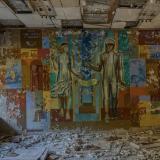 A chipping mural depicts a Soviet Kosmonaut and a woman of science on wall of a decaying building. The floor is littered with grey rubble and the ceiling tiles are cracked and falling.