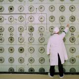 A nuclear plant worker in a white lab coat and medical hat adjust knobs on a pale green wall covered with number dials at the Windscale Pile One nuclear power plant.