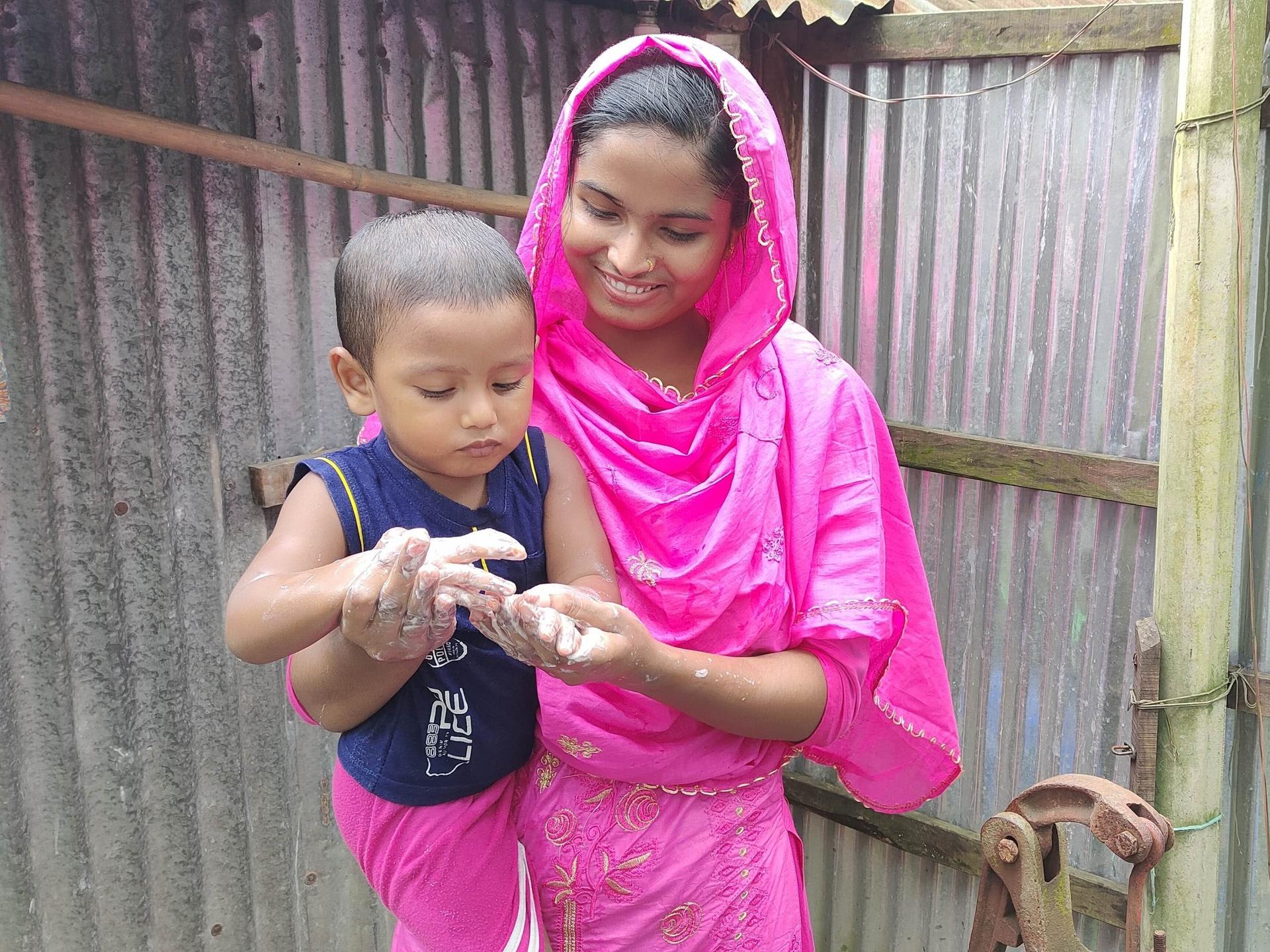 Health leader Sabikunnahar, wearing a bright pink traditional outfit, smiles while helping a young child wash their hands with soap and water outdoors beside a corrugated tin wall.