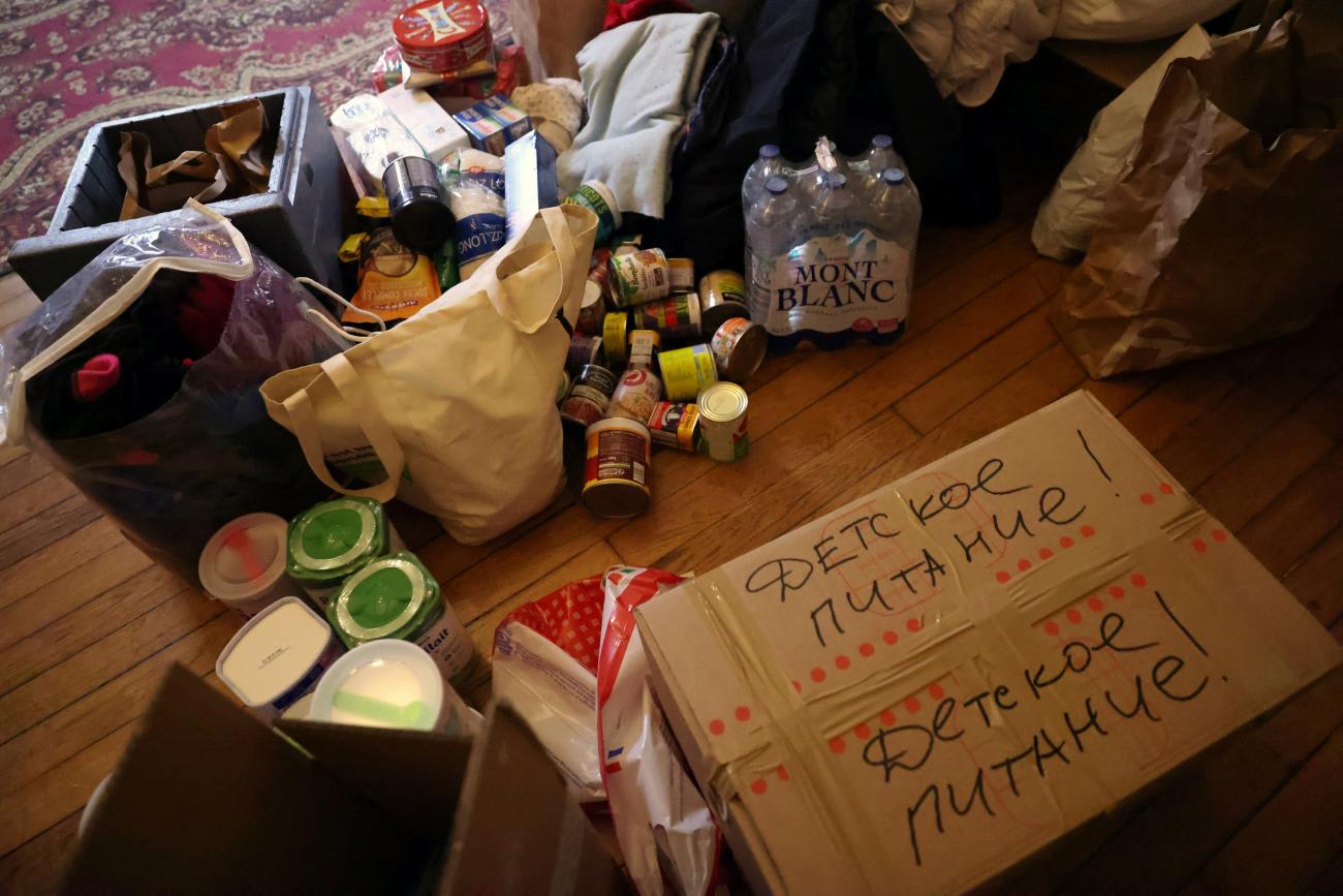 A cardboard box labeled "Baby Food" in Russian sits on a wood floor with an oriental rug surrounded by other canned goods and donated items.