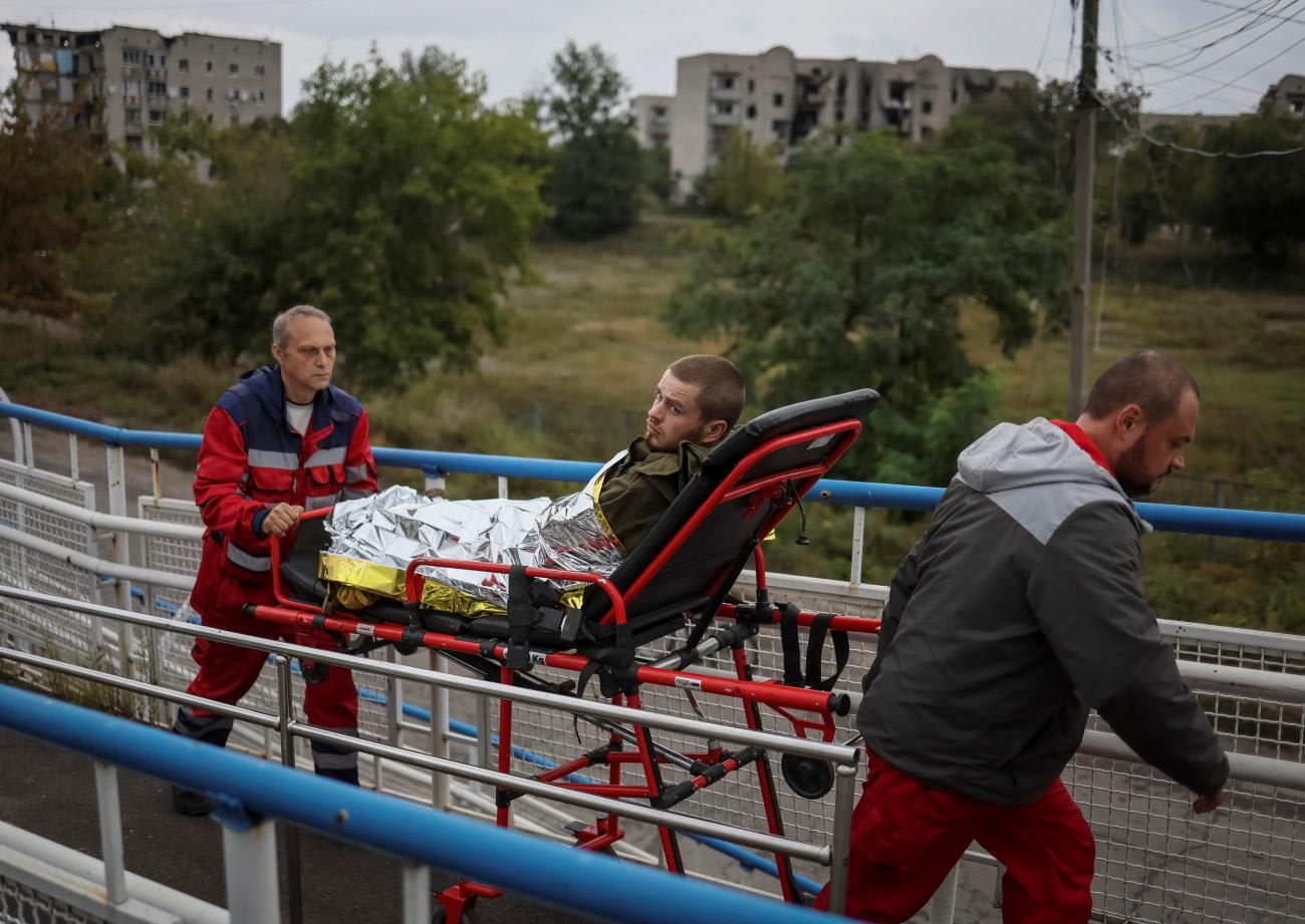 Two health workers in red emergency outfits transport a wounded Ukrainian serviceman on a stretcher in the town of Izyum, recently liberated by Ukrainian Armed Forces. In the background, are trees and concrete apartment buildings that have been damaged by shelling.