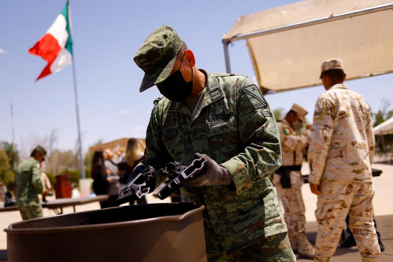In the center, a Mexican soldier in a green camouflage uniform and black mask puts pieces of a dismantled gun in a bin. Behind him a Mexican flag flies over other soldiers in khaki uniforms working under a beige tent.