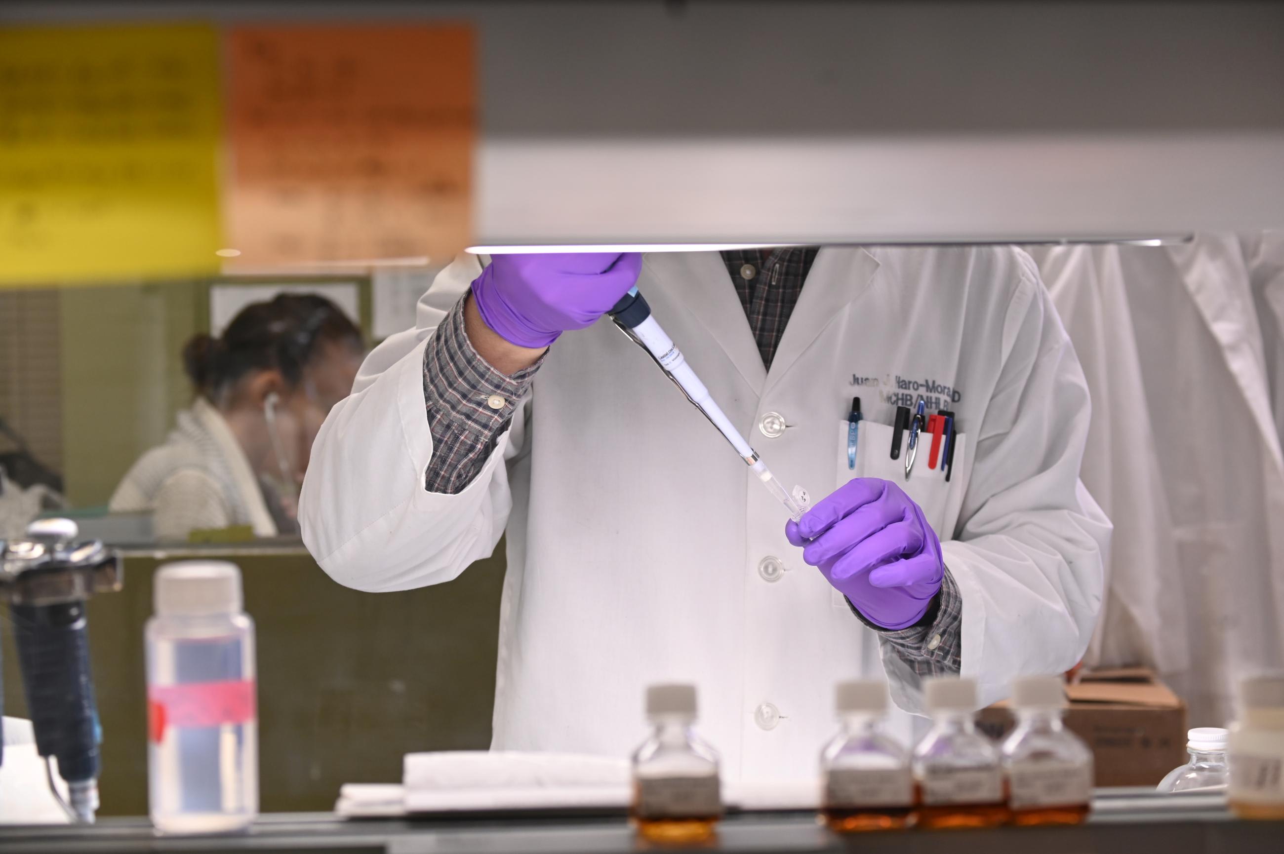 A researcher, wearing a white lab coat, whose face is obscured by white cabinetry, extracts liquid via syringe from a test tube while wearing purple latex gloves