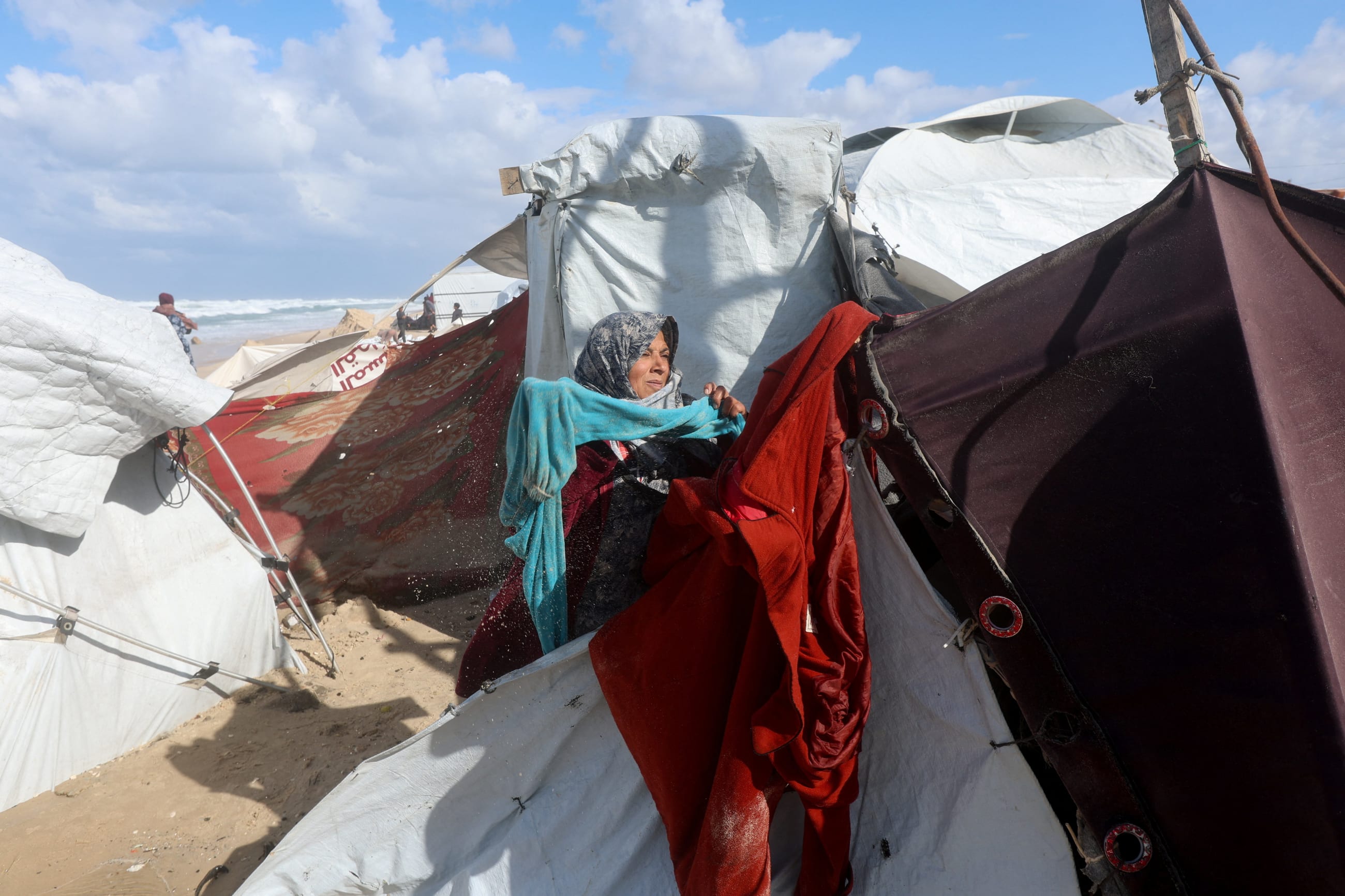 A displaced Palestinian woman collects wet clothes at a beach tent camp after it was flooded by rising seawater during a winter storm, in Khan Younis, southern Gaza Strip, on December 28, 2025.