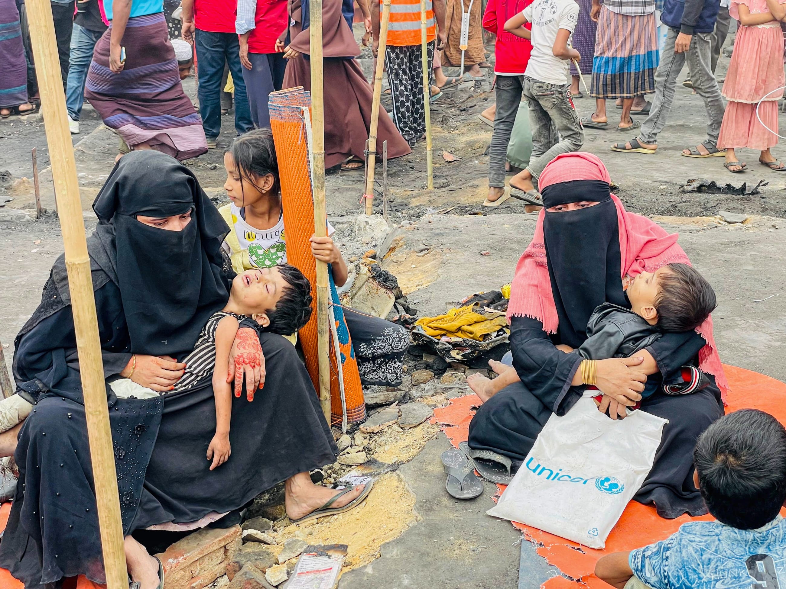 Two women cradle their children in the ruins of a burned section of the encampment, where swaths of homes were reduced to ash, in Cox's Bazar, Bangladesh, on December 25, 2024.