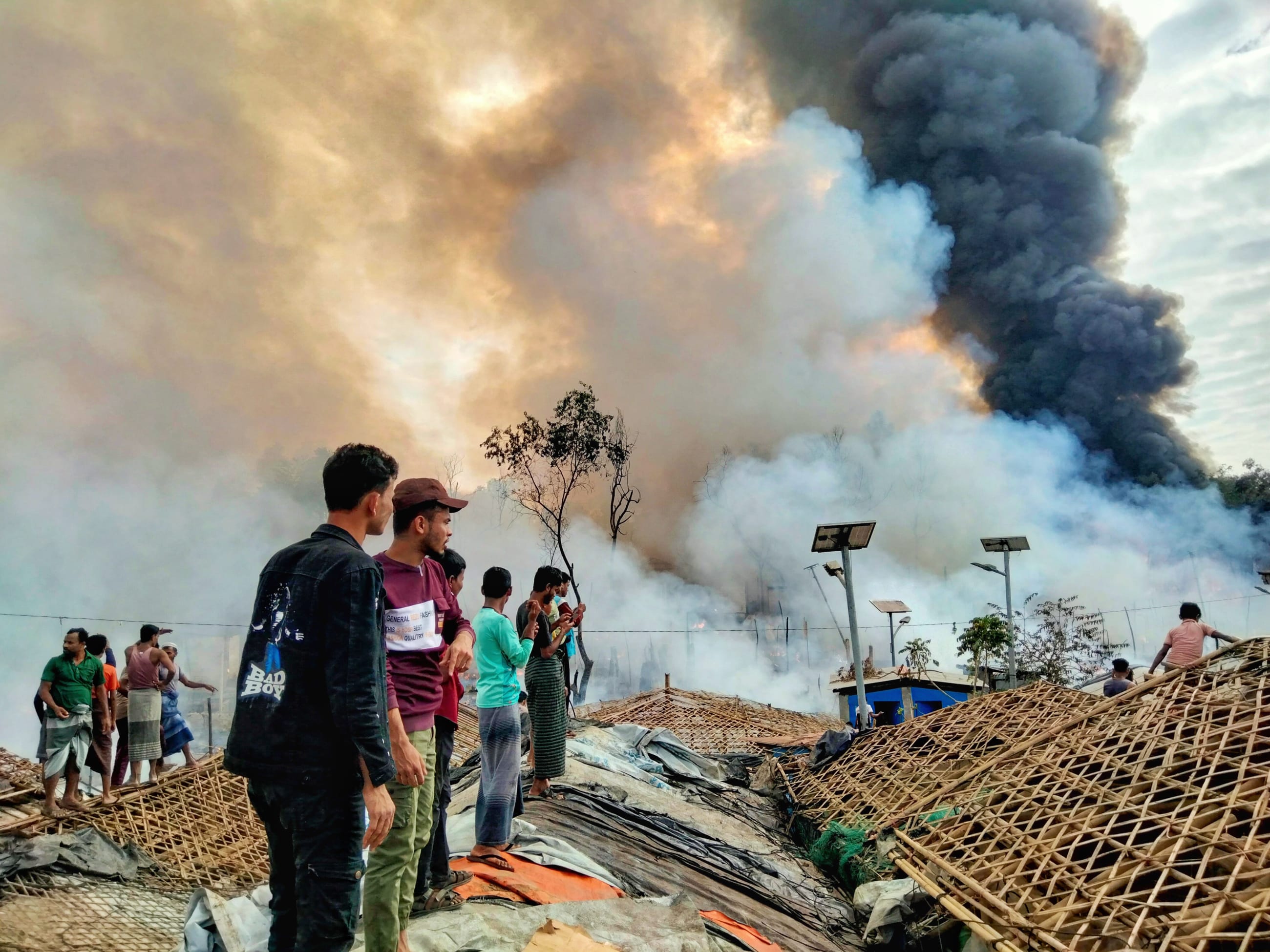 Young men stand on bamboo roofs of the remaining shelter as a fast-moving fire consumes large sections of the camp, in Cox's Bazar, Bangladesh, on December 24, 2024.