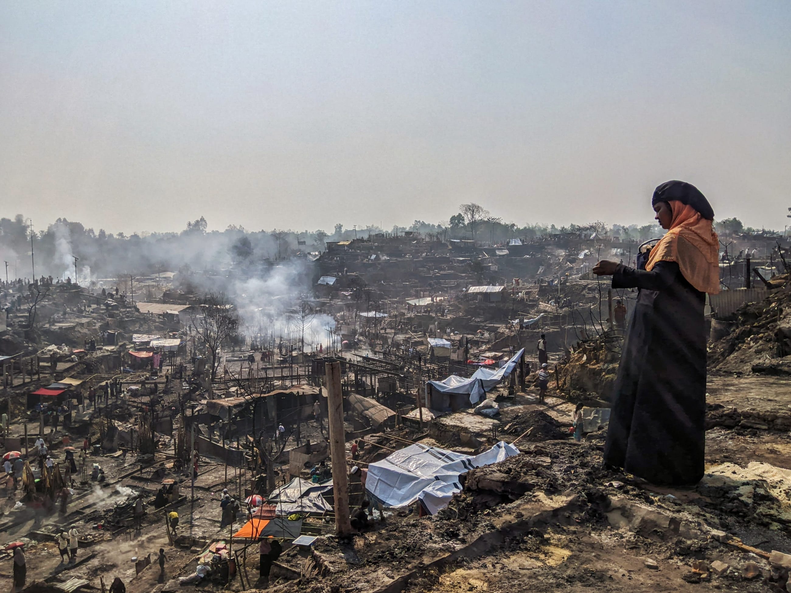 A Rohingya woman overlooks the charred remains of her camp as smoke rises and survivors sift through what little is left, in Cox's Bazar, Bangladesh, on March 6, 2023.