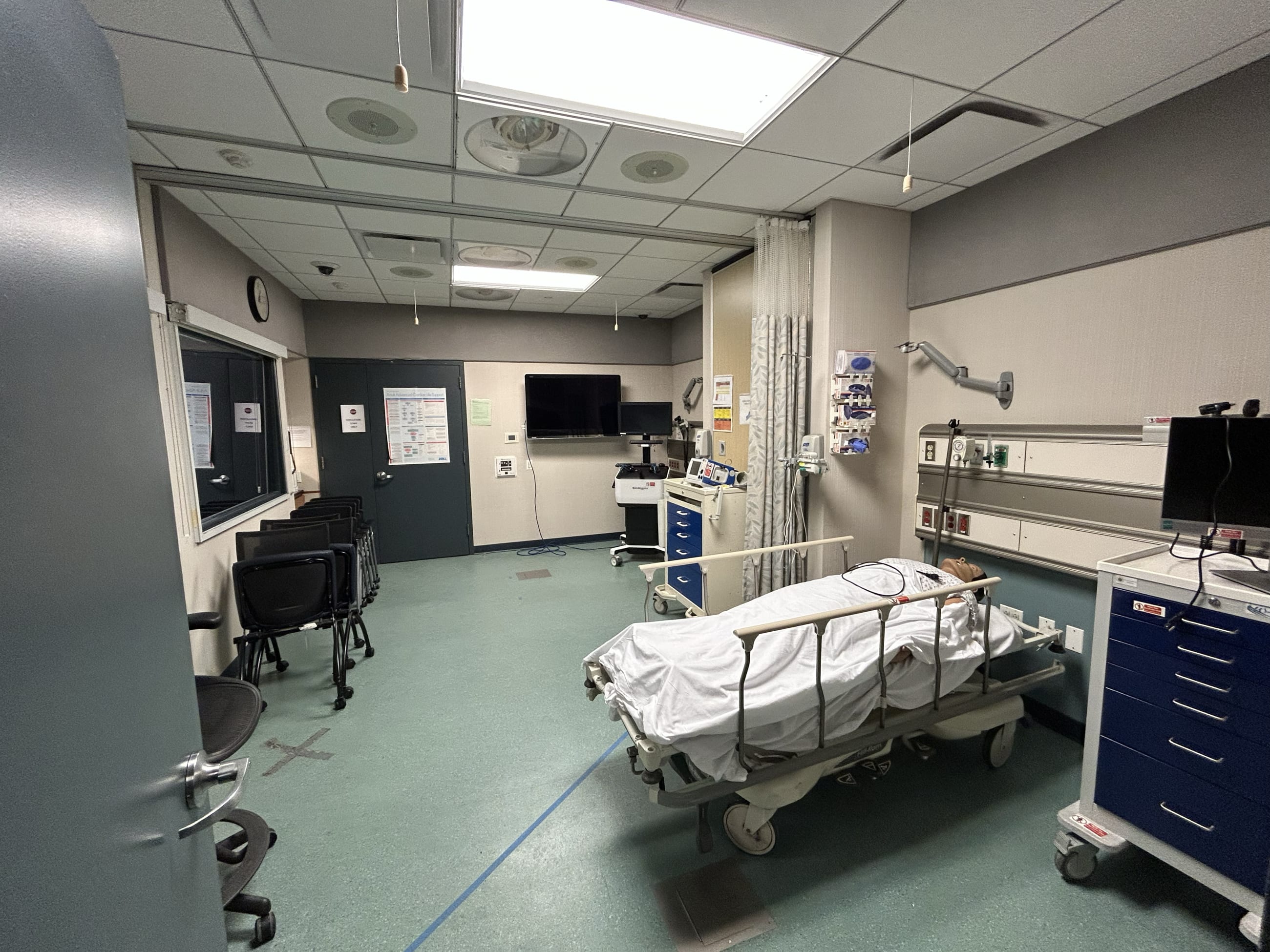 A clinical simulation room at the Institute of Medical Simulation and Learning, where a patient mannequin lies in a fully equipped mock hospital bay. The one way observation window at left allows instructors to watch trainees without interfering, on February 2, 2026.