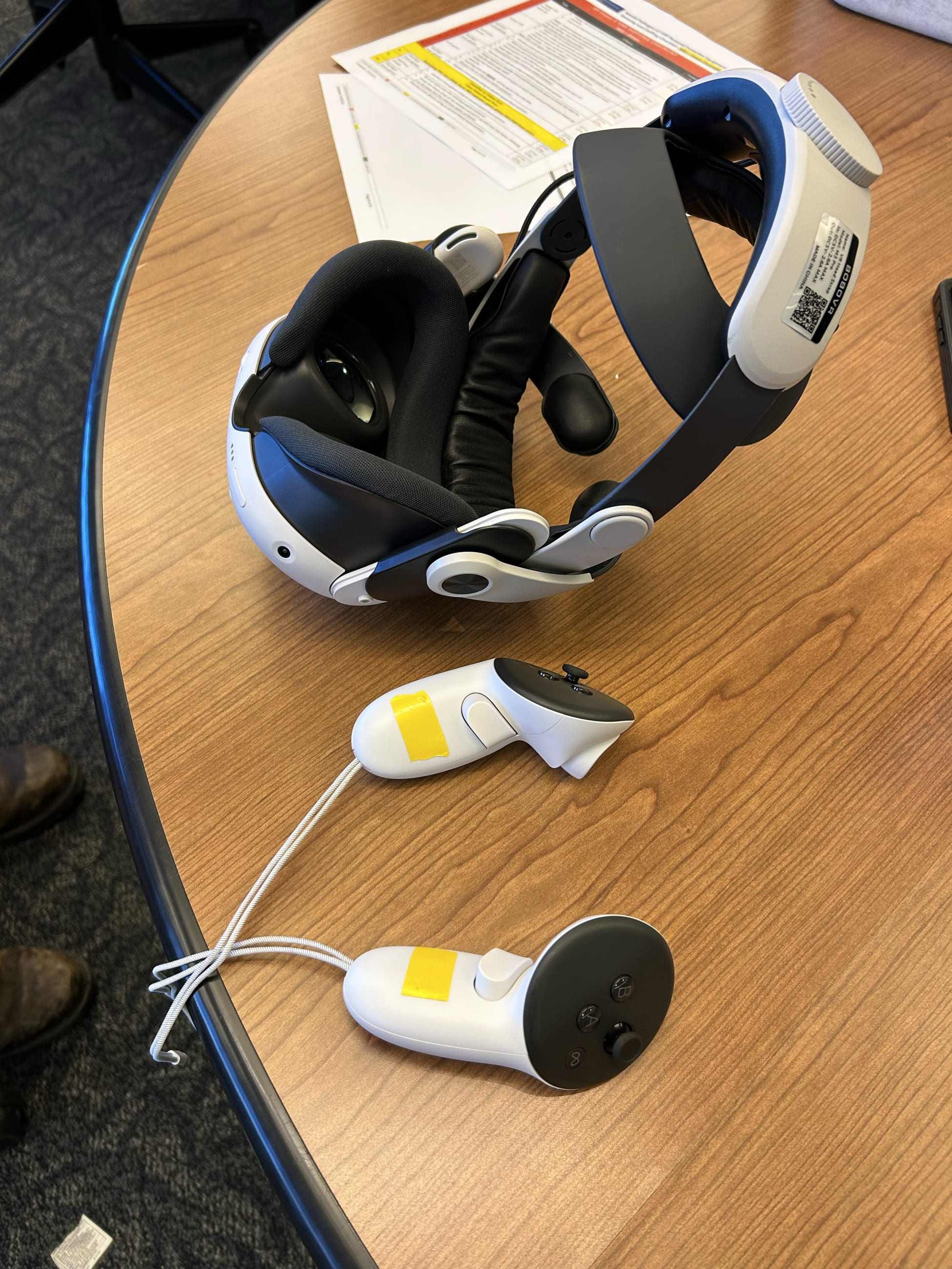 A virtual reality headset and paired hand controllers sit on a table at NYC Health + Hospitals' Institute of Medical Simulation and Advanced Learning at the Jacobi Medical Center in the Bronx, on February 2, 2026. The yellow color-coded checklist for personal protective equipment (visible in the background) corresponds to the "yellow zone" doffing protocol the VR module is designed to teach.