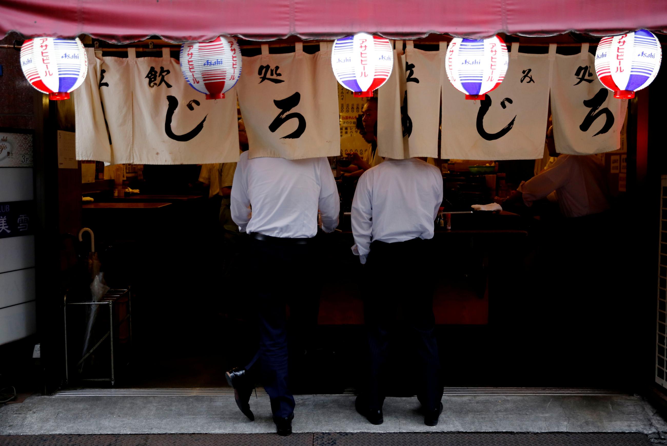 Two men wearing white shirts and black slacks stand with their backs facing the camera and their heads obscured by beige signage with black Japanese characters