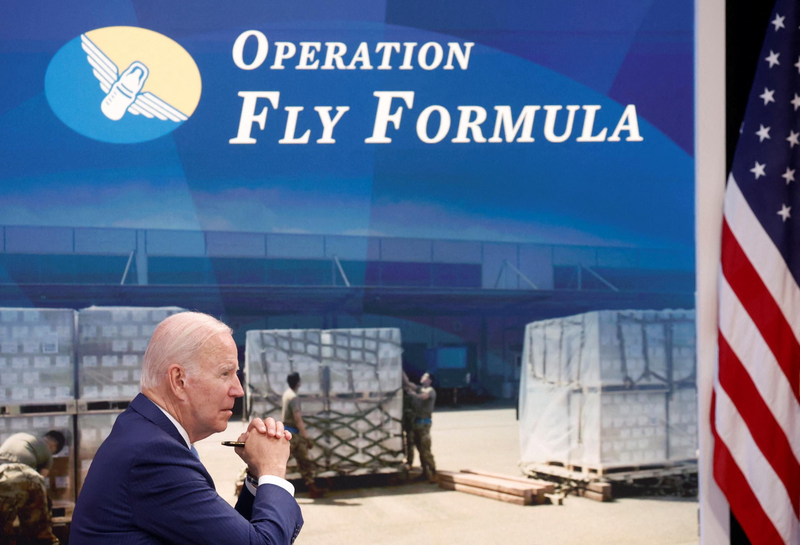 President Joe Biden sits, looking pensive, facing an American flag. Behind him is a screen depicting U.S. soldiers unloading crates of formula beneath a sign that says "Operation Fly Formula" next to a blue and ivory logo of a baby bottle with wings.