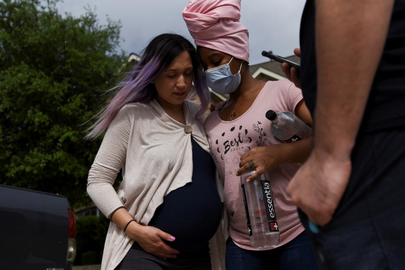 Nancy Pedroza, 27, who is pregnant and wearing a white sweater, is supported by her Nichollette Jones, her doula, who is wearing a pink head scarf and medical mask, as they take a walk to help speed up Pedroza's contractions in Fort Worth, Texas, United States, on April 7, 2020.