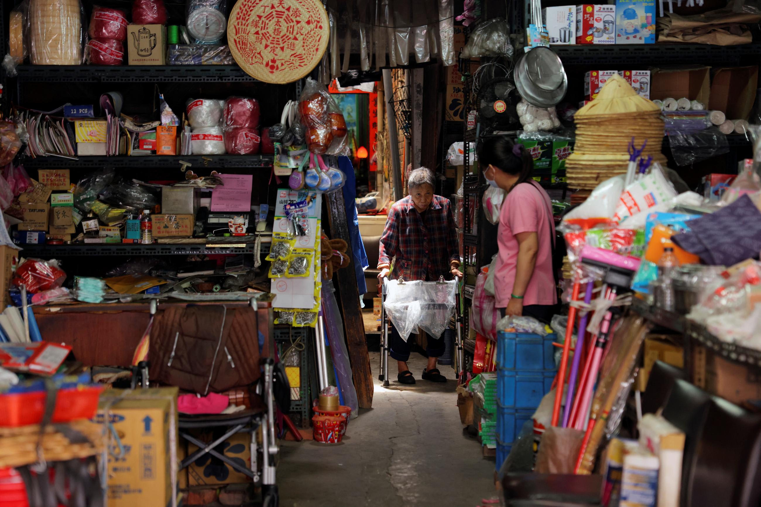 An elderly woman walks with a walker through a doorframe in the back of a hardware store, on either side of which are shelves laden with goods in New Taipei, Taiwan on April 22, 2022.