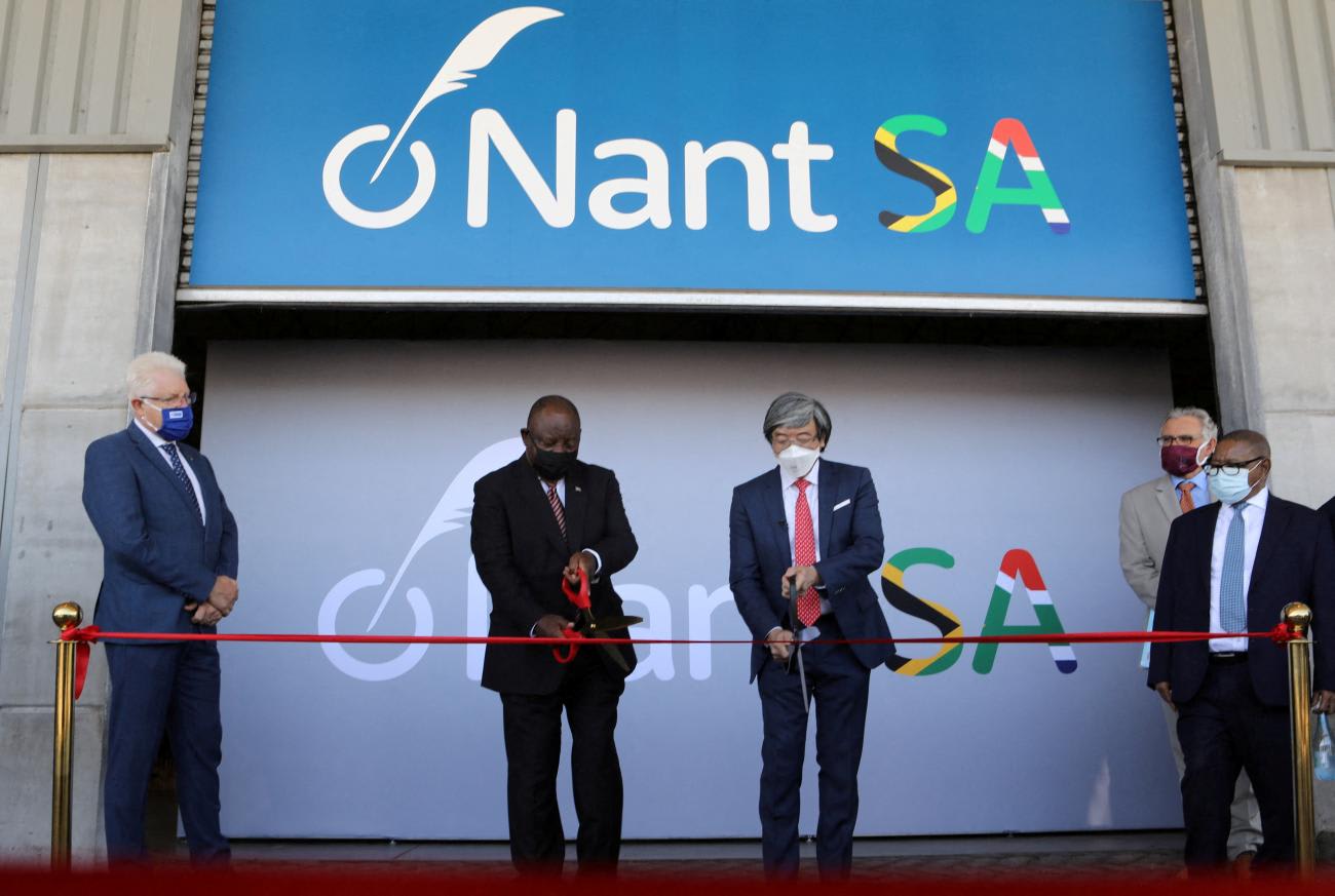 South African President Cyril Ramaphosa wears a black suit as he cuts a ribbon in front of a blue background with the NantSA logo during the launch of NantSA, a future vaccine manufacturing facility designed to accelerate the production of pharmaceuticals, in Cape Town, South Africa, on January 19, 2022.