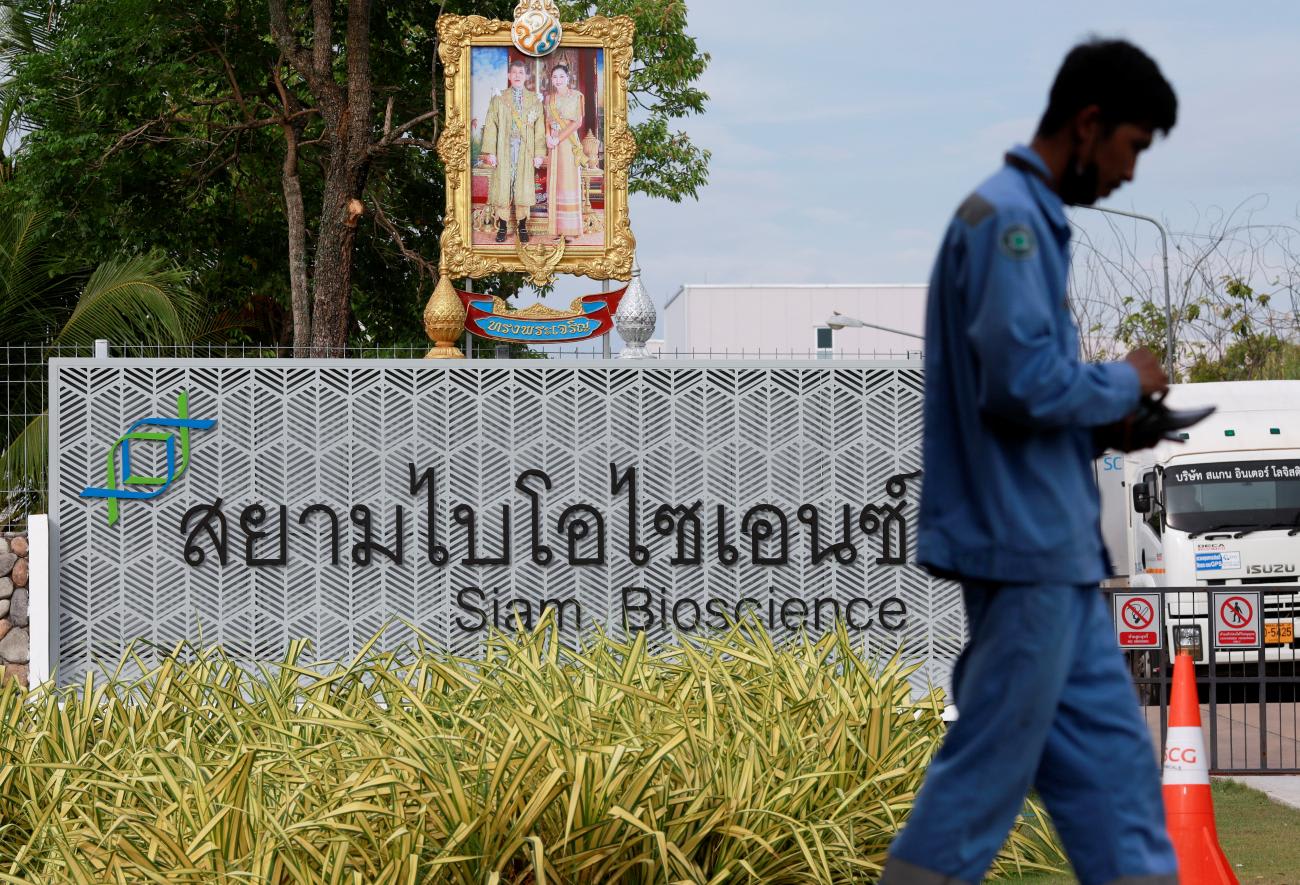 A man walks in front of a sign for the Siam Bioscience factory, where the vaccine against COVID-19 will be produced. The sign is topped with a portrait of the Thai royal family in a gold frame, and surrounded by tall manicured grass. The photo was taken outside Bangkok, Thailand on June 3, 2021.