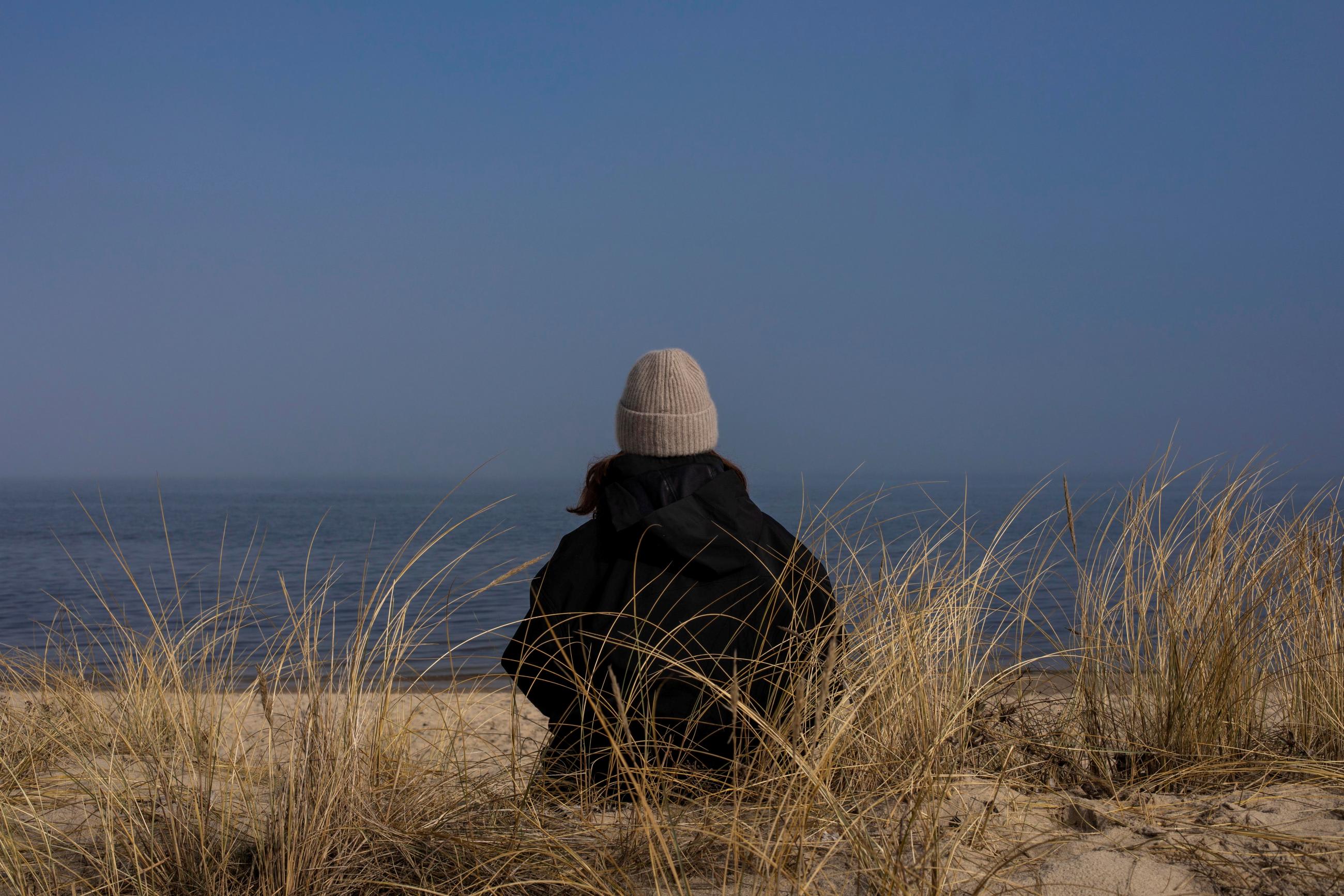 A woman in a black coat and gray hat is seen from behind as she gazes out at the sea while sitting on a sandy beach surrounded by tall grass in the evening.