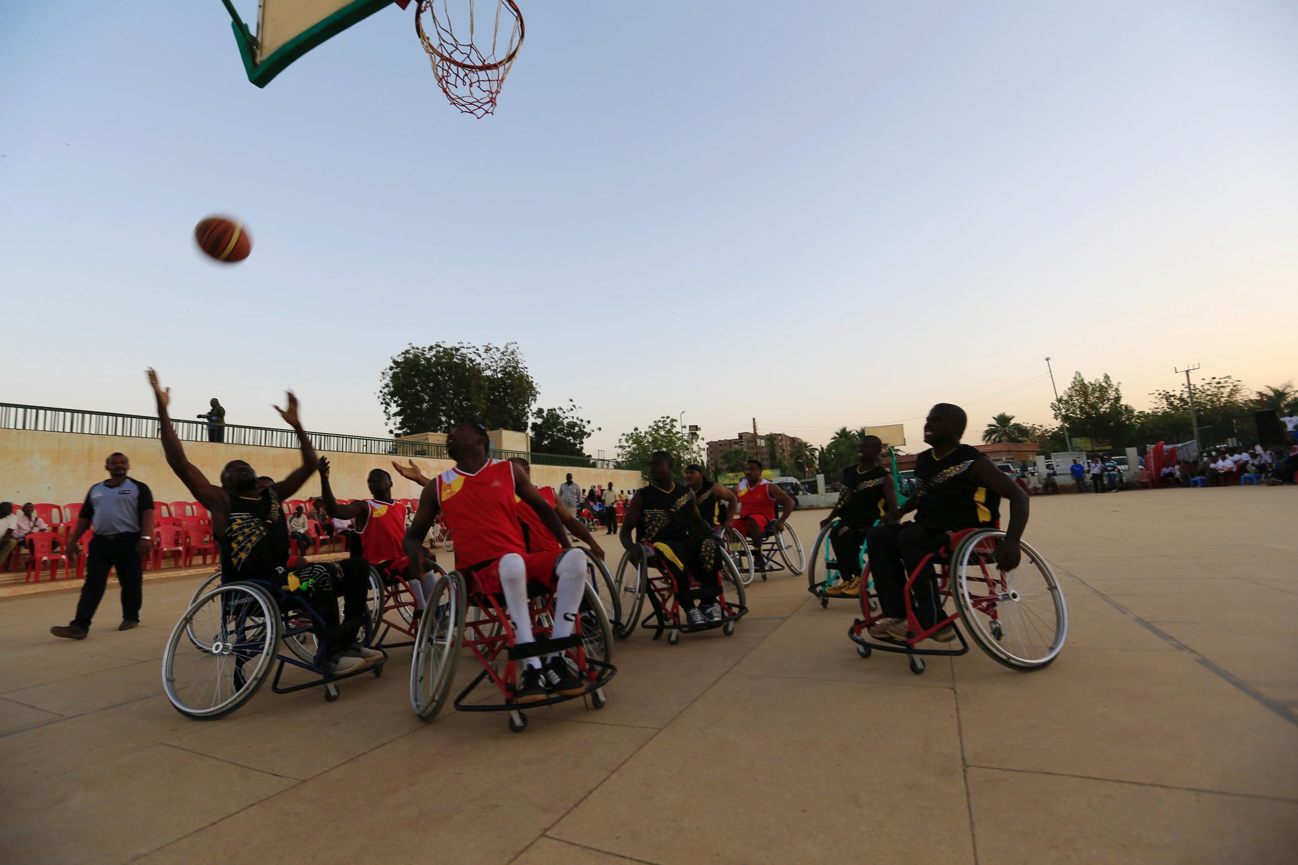Two teams, one in red jerseys, the other in black jerseys, face off during the first wheelchair basketball tournament in Sudan, marking the International Day of Persons with Disabilities at dusk.